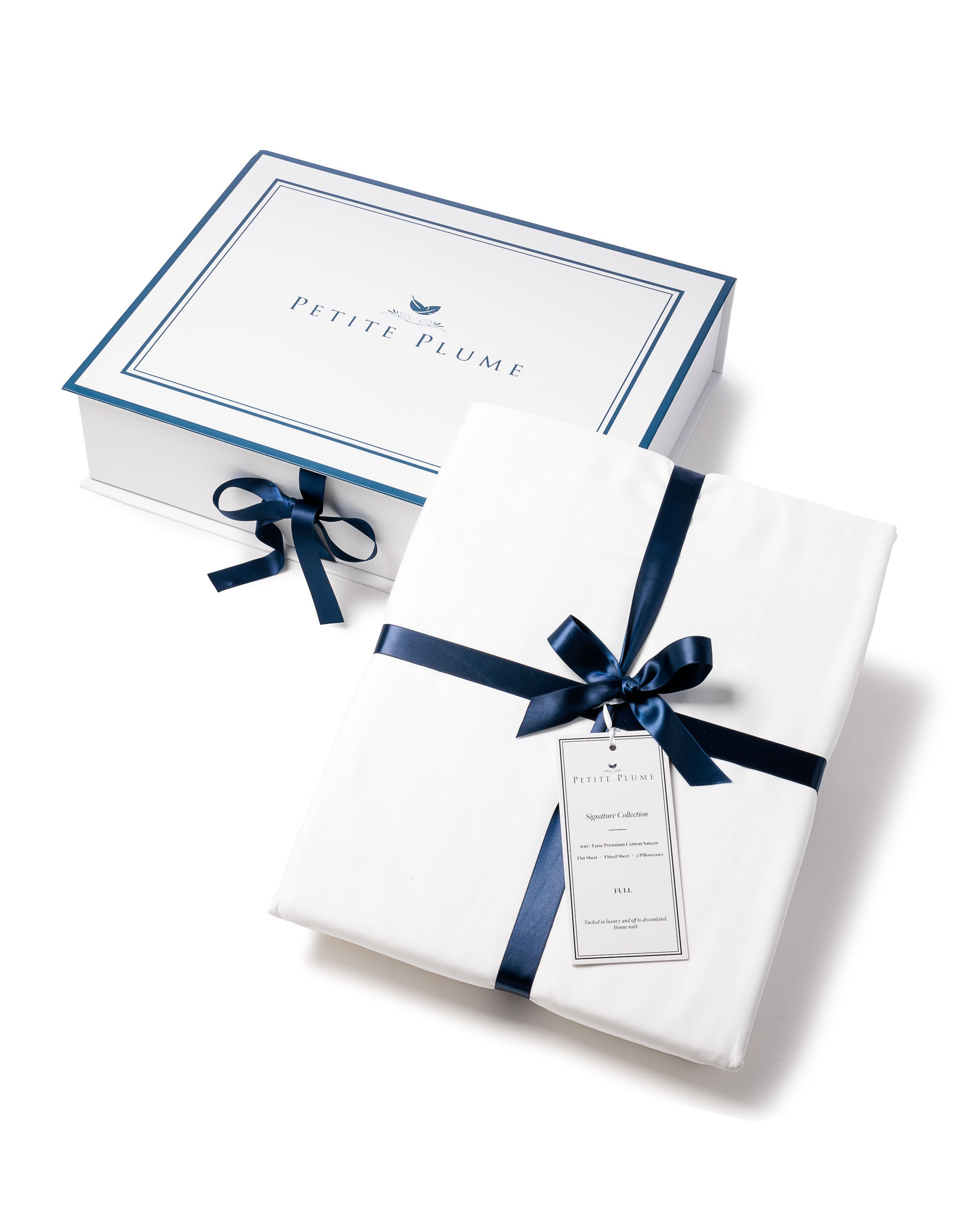 A Petite Plume packaging box in white is adorned with a blue ribbon and bow. Beside it lies Signature Luxe Sateen Bed Sheets in white, made from 100% long-staple cotton, elegantly tied with a matching blue ribbon and tag, echoing five-star hotel presentation.