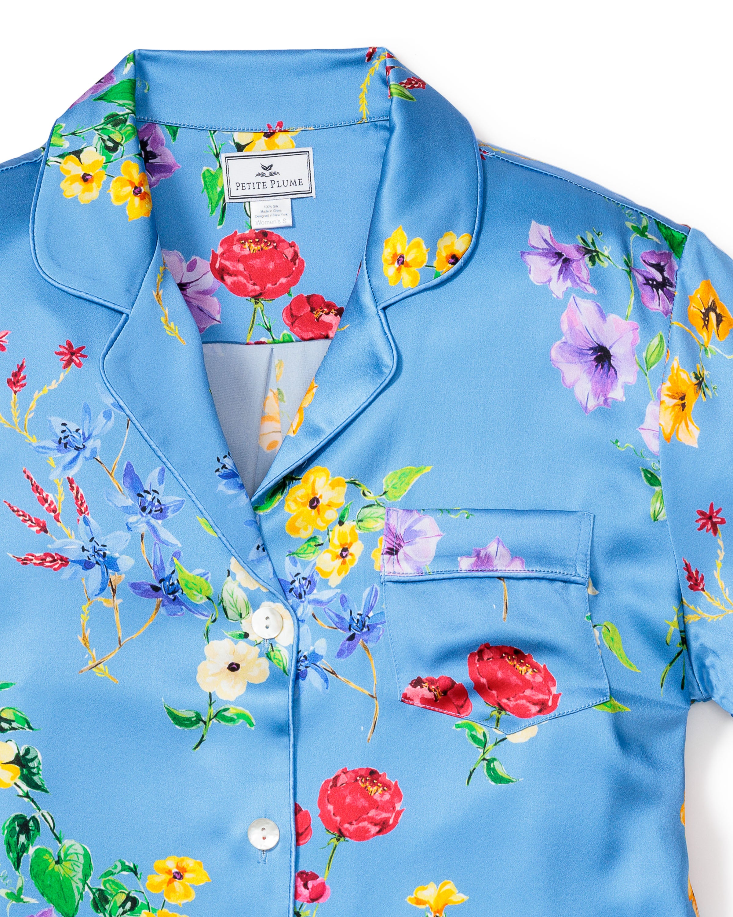 The Womens Silk Pajama Set in Brilliant Botanical Azure by Petite Plume features a blue top with a vibrant botanical print, showcasing red, yellow, purple, and blue flowers. This luxurious shirt includes a collar, white buttons, and a chest pocket.