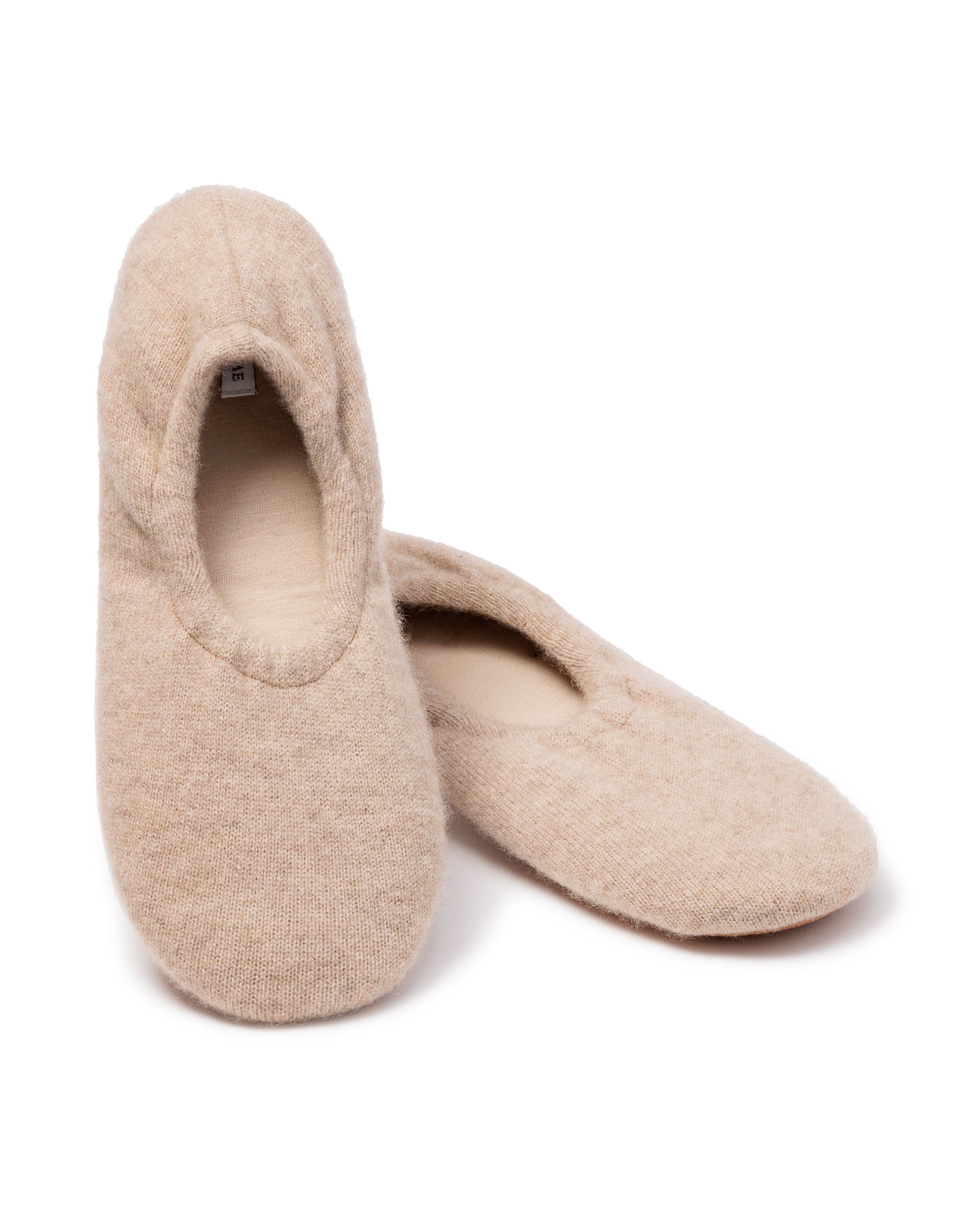 A pair of Petite Plume Womens Cashmere Slippers in Beige, featuring a simple, soft design. One slipper lies flat while the other is slightly propped up behind it, highlighting the cozy fit and rounded opening.