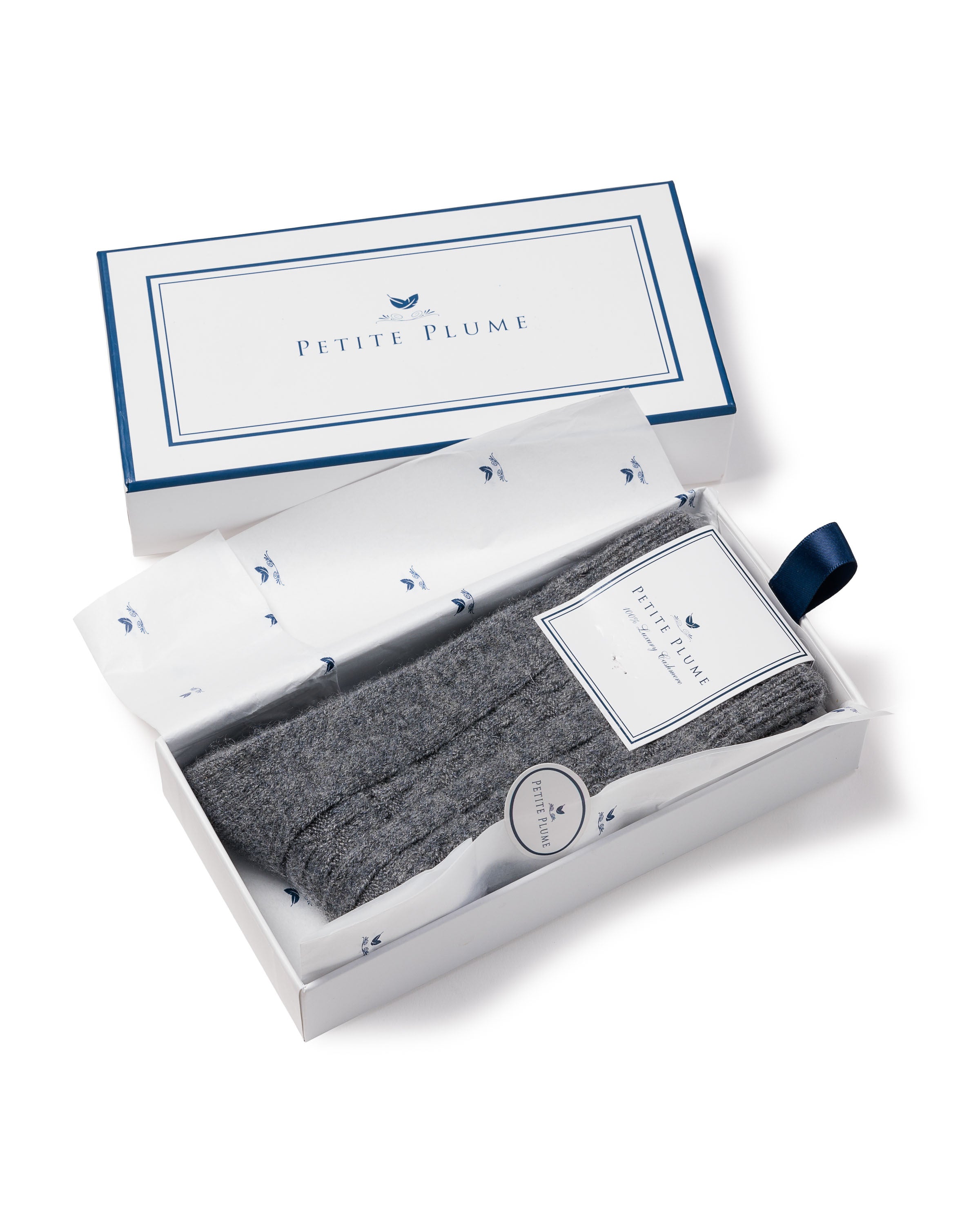 An open box reveals folded Mens Cashmere Socks in Dark Grey on white tissue paper, exuding luxurious comfort. The tag reads “Petite Plume.” Nearby, the cover features a blue outline and a small blue feather icon, suggesting the elegance within this gift box.