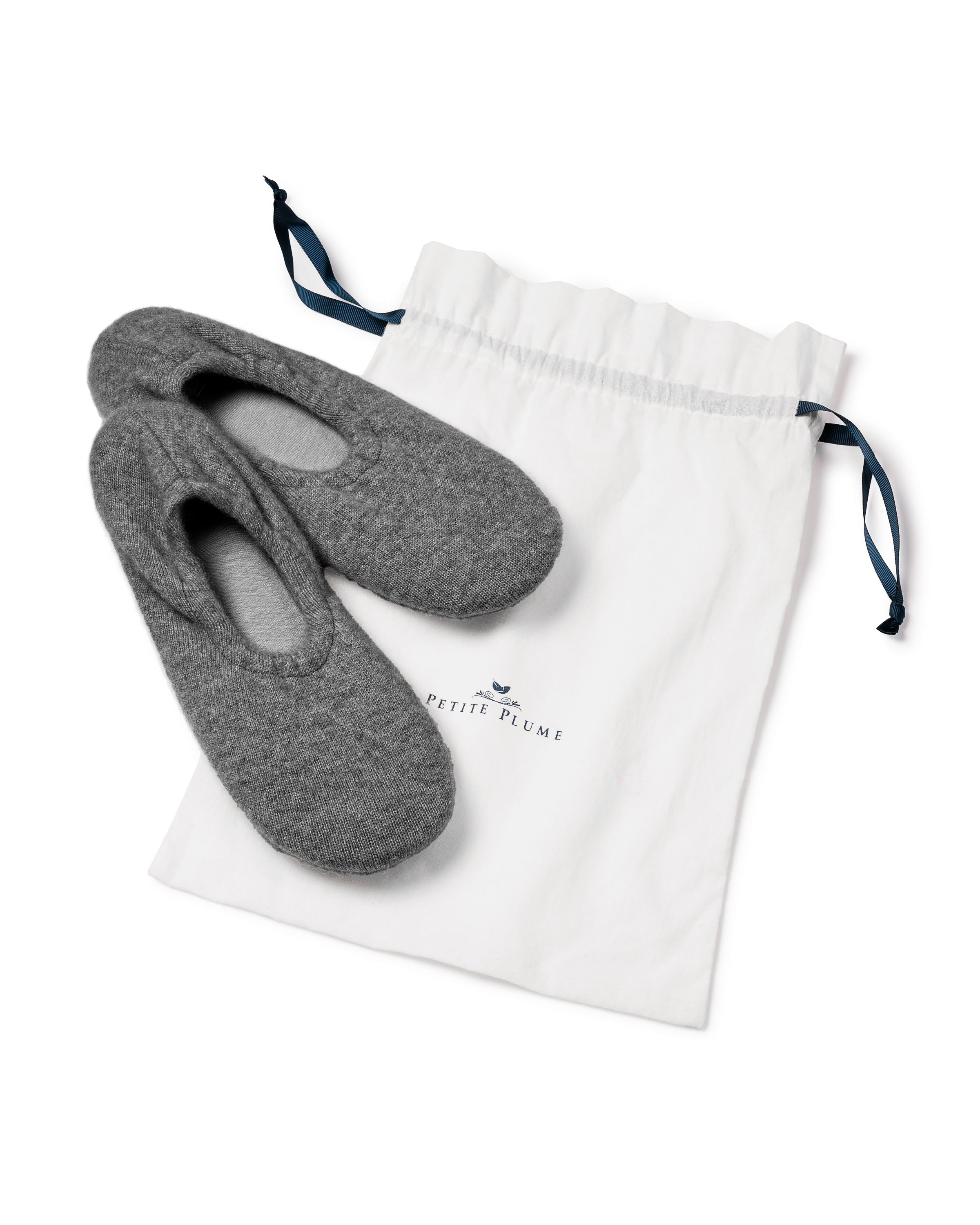 Womens Cashmere Slippers in Dark Grey, skillfully designed for comfort, are displayed on a white drawstring bag with blue ties. The bag features Petite Plume and a small bird logo, embodying a cozy, minimalist style.