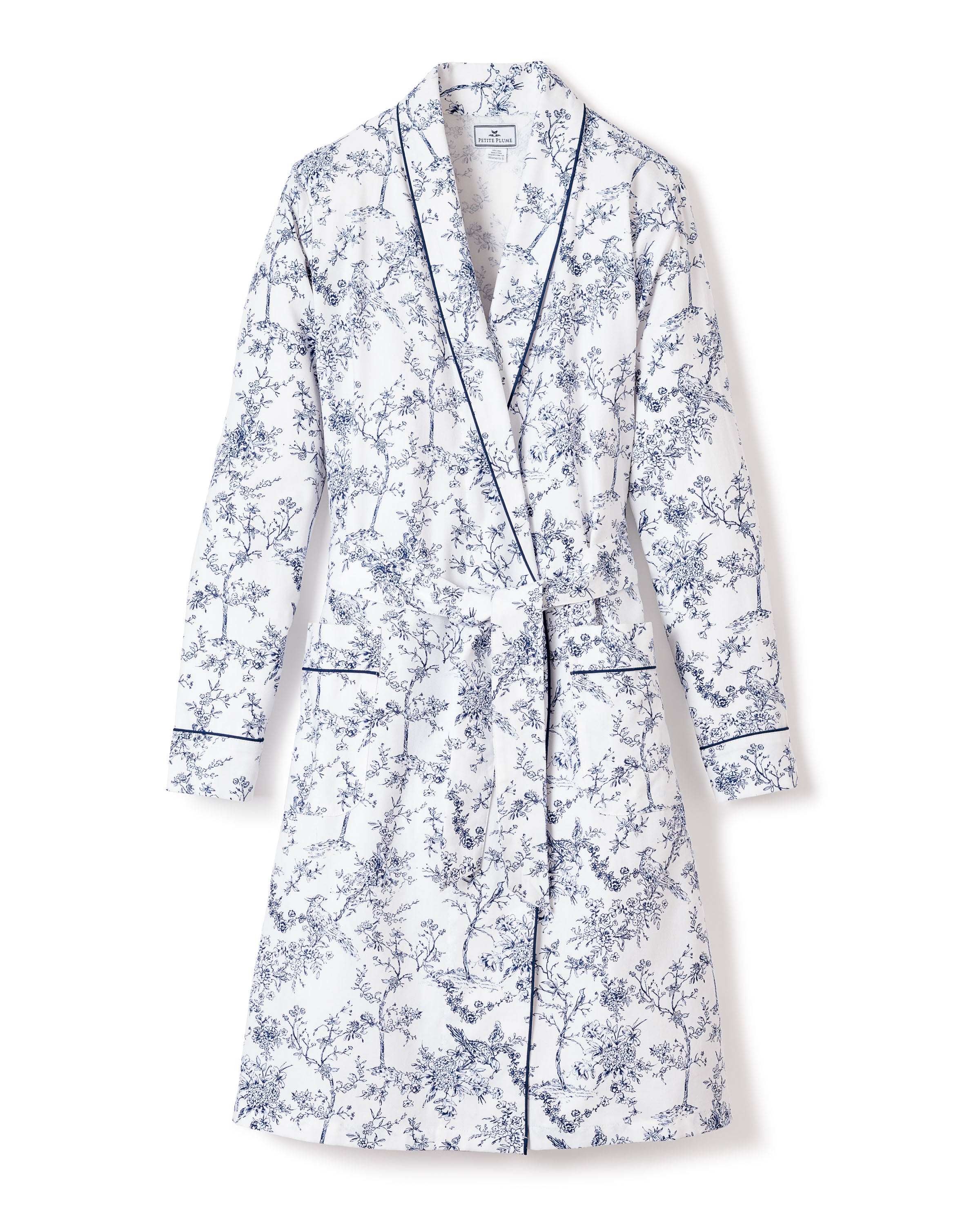 The Womens Twill Robe in Timeless Toile by Petite Plume, crafted from the finest quality cotton, features a white and blue floral design with a shawl collar, long sleeves, belted waist, and refined blue piping on the edges—exuding botanical elegance against a plain backdrop.