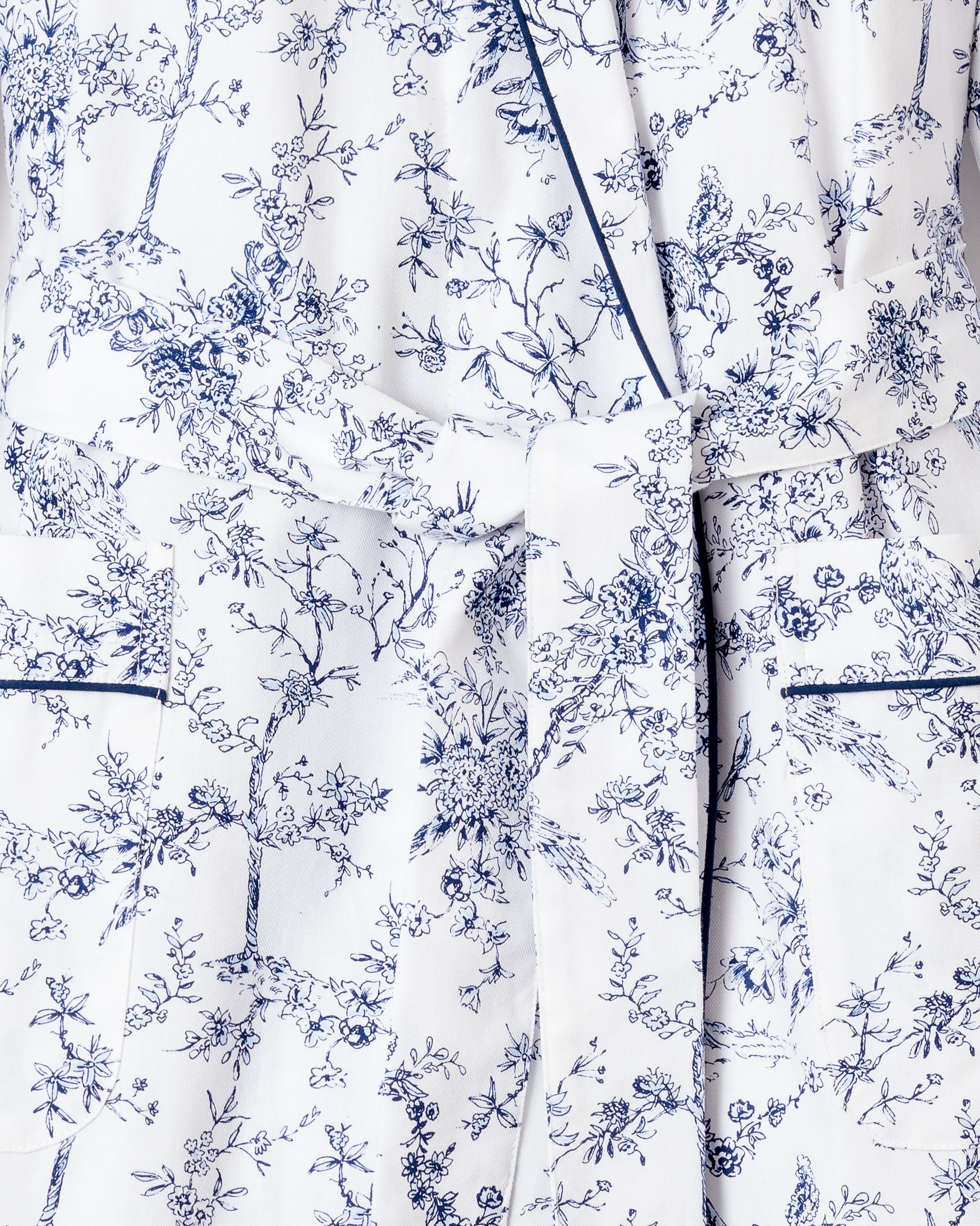 A close-up of the Womens Twill Robe in Timeless Toile by Petite Plume reveals a detailed blue and white toile de Jouy pattern. Made from high-quality cotton, the robe includes a belt and pockets, exuding botanical elegance with intricate floral and pastoral designs.