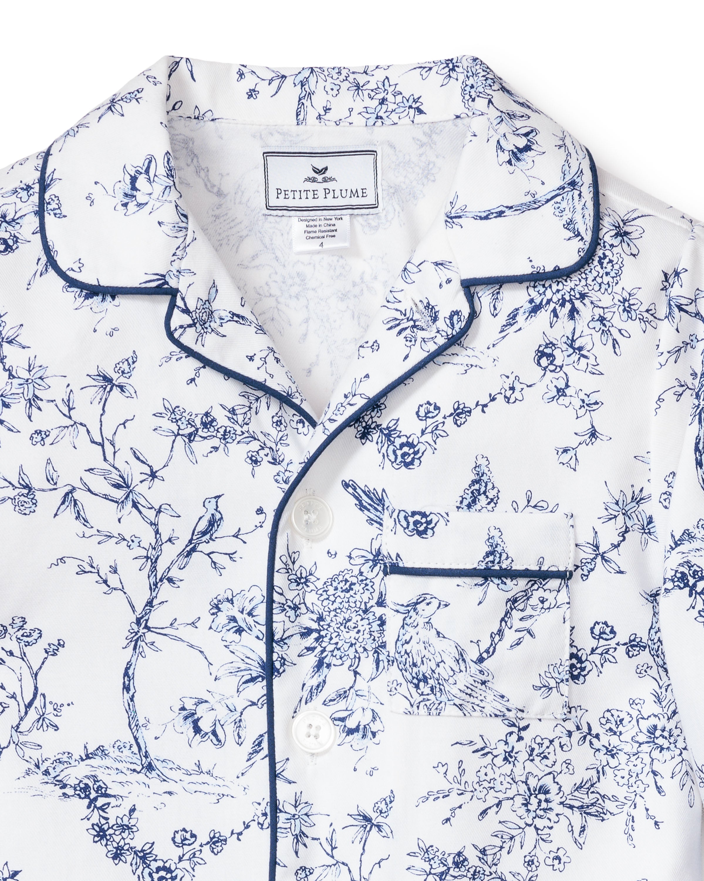 The Kids Twill Pajama Set in Timeless Toile by Petite Plume showcases botanical elegance with its close-up of a white top adorned with blue floral and bird prints. Featuring a classic collar, front buttons, and a chest pocket with contrasting blue piping, it is crafted from luxury sleepwear material.