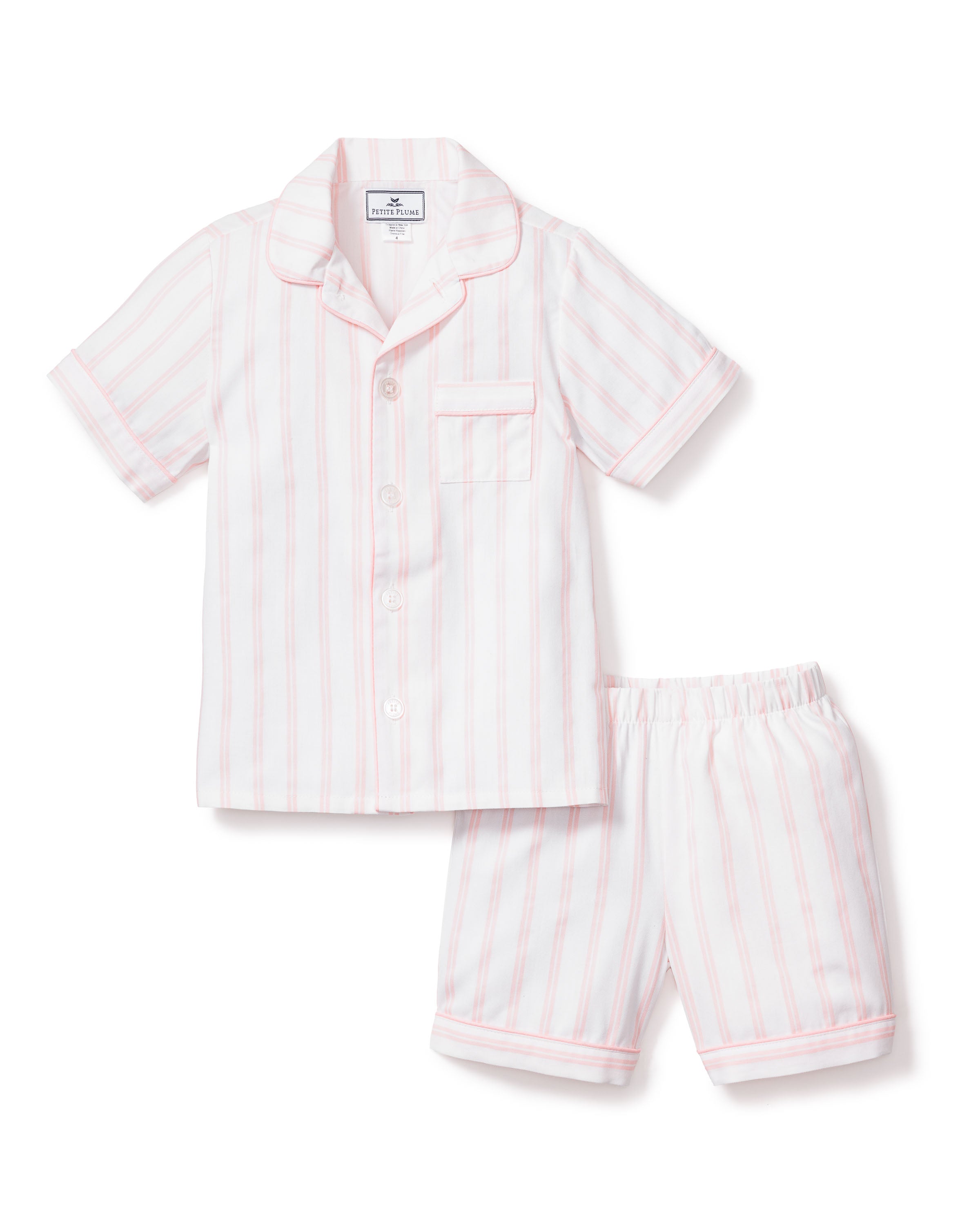 The Kids Twill Pajama Short Set by Petite Plume features a luxurious cotton feel with white short sleeves and pink stripes. The collared top includes buttons and a front pocket, while the shorts offer an elastic waistband for comfort.