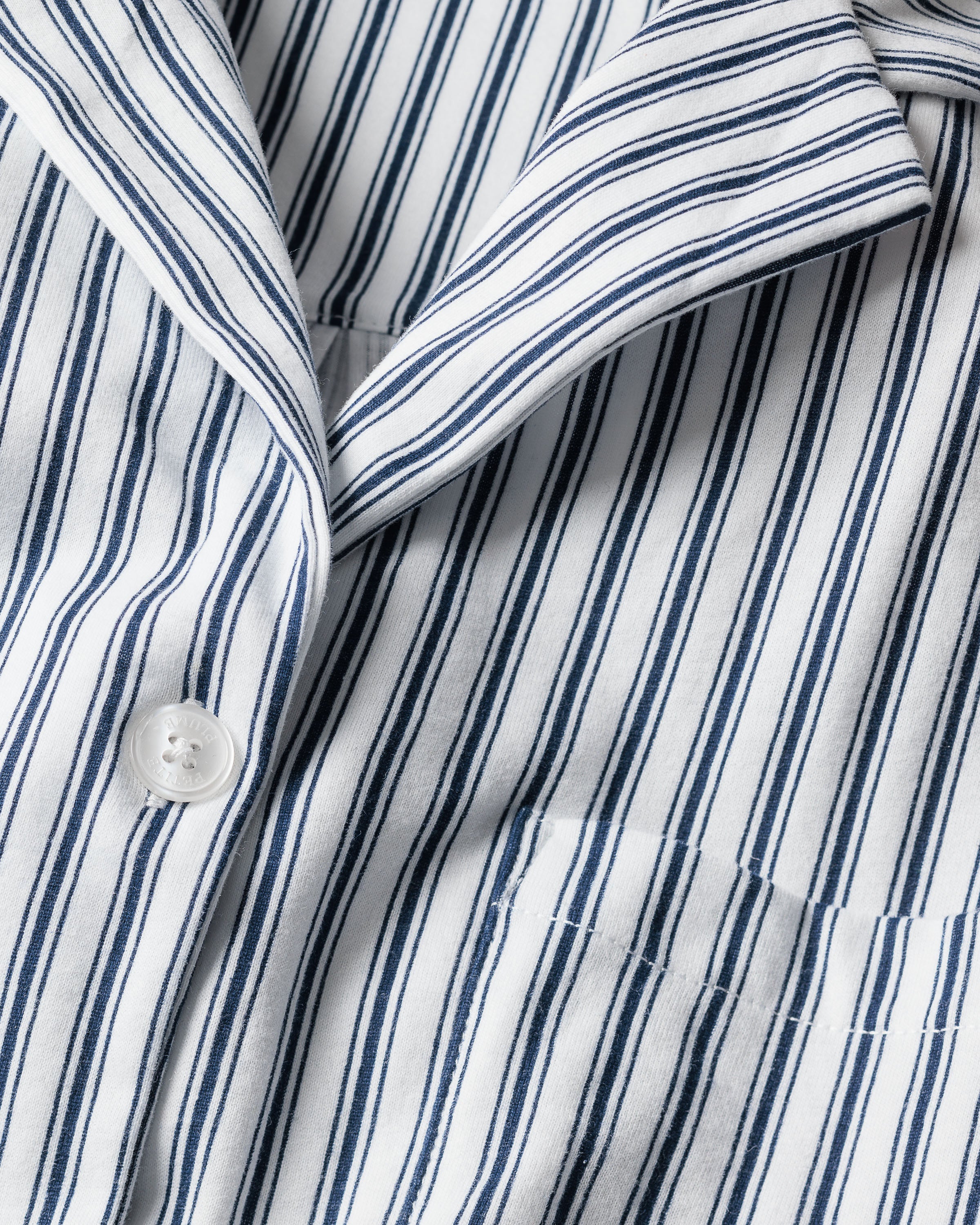 A close-up of The Olivia Wide Leg Pima Pajama Set in Navy French Ticking by Petite Plume showcases a button and pocket on vertical navy blue and white stripes, with the rich texture of Peruvian Pima cotton offering a classic, tailored design.