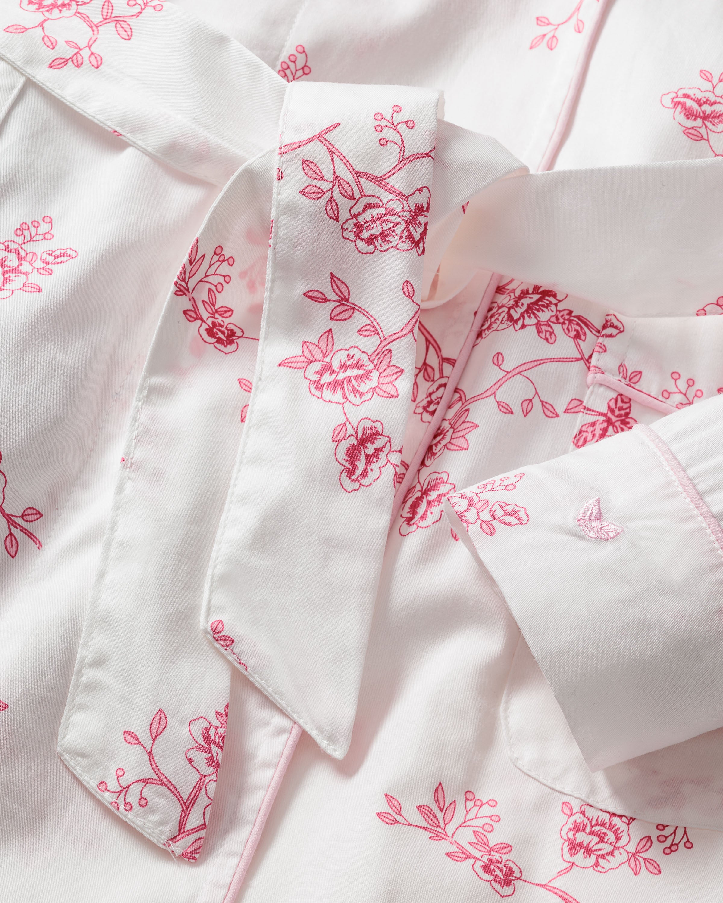 Close-up of a white fabric with pink floral embroidery hints at a vintage print. The collar and fabric tie suggest its the Womens Twill Robe in English Rose Floral from Petite Plume, crafted from high-quality cotton.