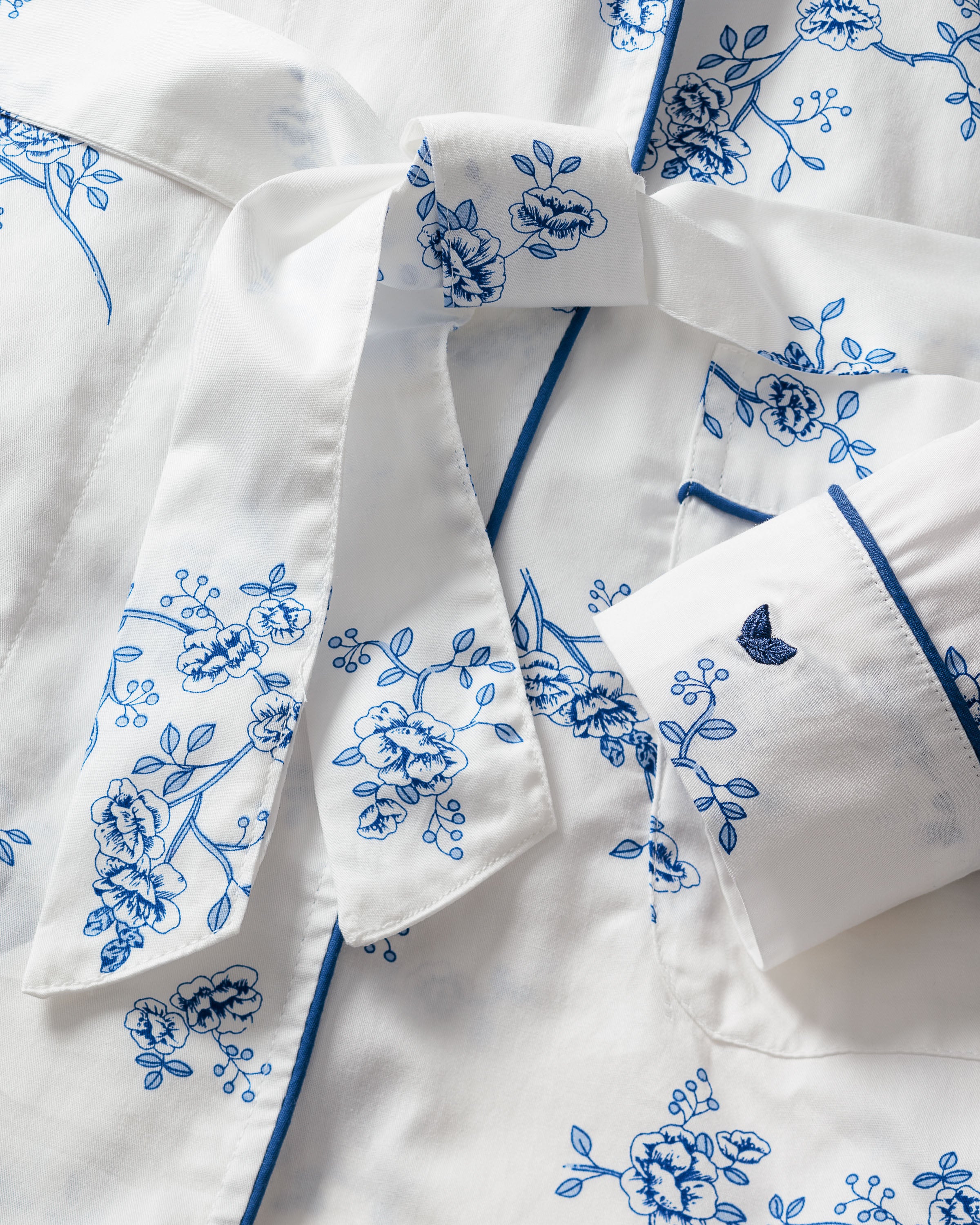 A close-up of the Womens Twill Robe in Indigo Floral by Petite Plume shows a white fabric with blue floral patterns, neatly folded sections, buttons, and ribbon-like accents. Crafted from premium cotton, it features stylized flowers and an embroidered butterfly on a cuff for vintage charm.