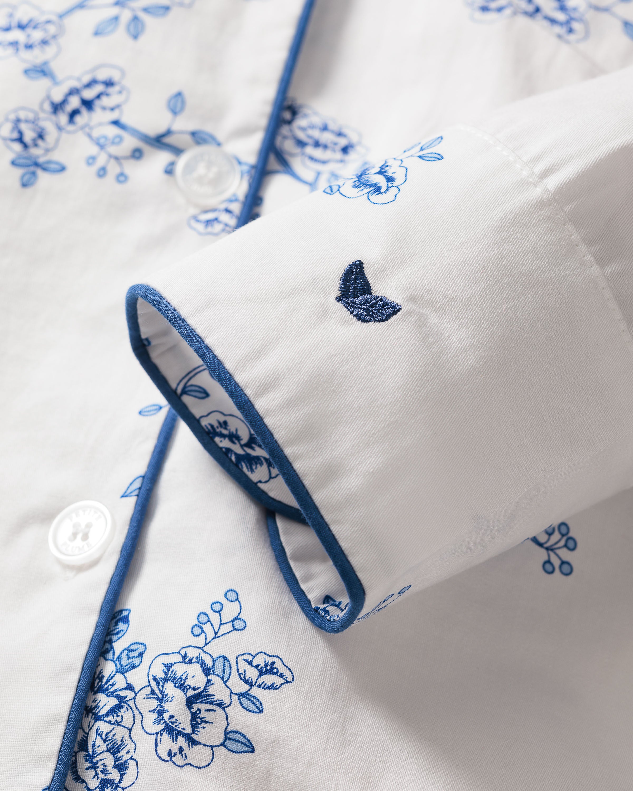 A close-up of the luxurious Womens Twill Pajama Set in Indigo Floral by Petite Plume shows a white shirt with blue floral patterns and dark blue piping. Made from the finest quality cotton, it features white buttons and an embroidered dark blue butterfly on the cuff for elegance.