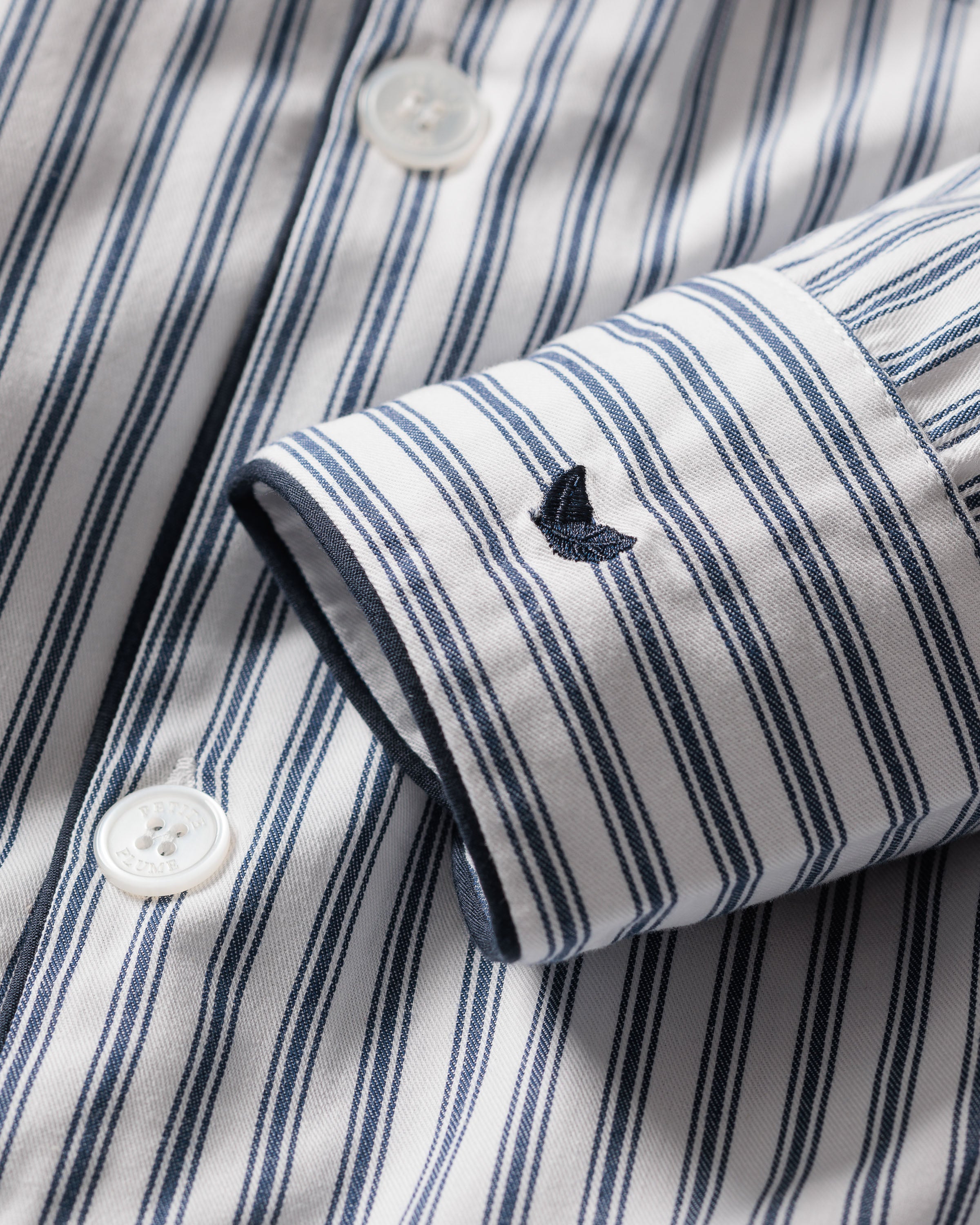 A close-up of Petite Plumes Kids Twill Pajama Set in Navy French Ticking shows a yarn-dyed cotton shirt with vertical blue stripes and an embroidered blue whale on the cuff, featuring white buttons.