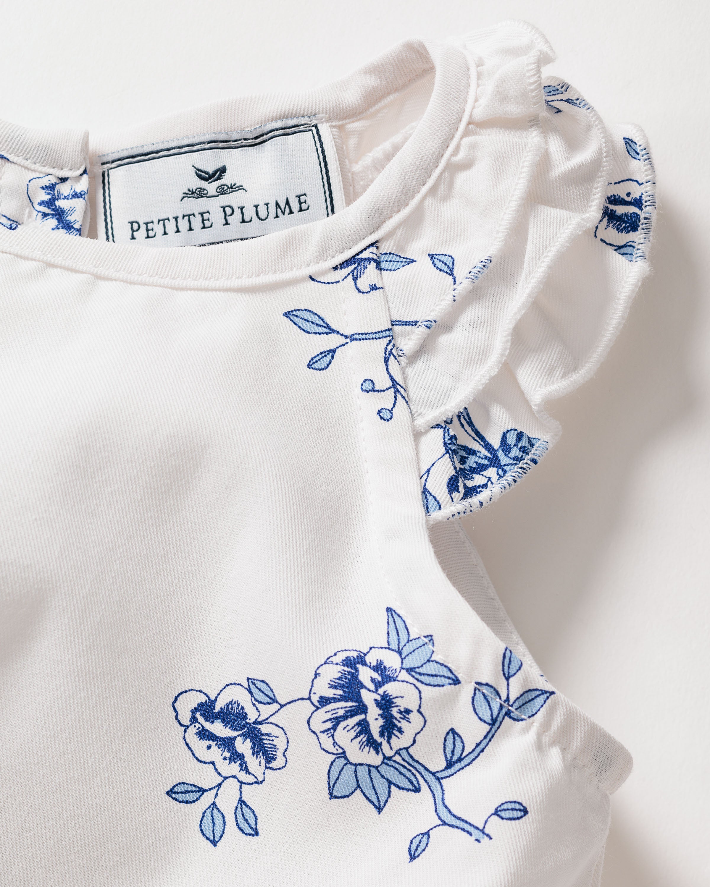 A close-up showcases the Girls Twill Amelie Nightgown in Indigo Floral by Petite Plume, highlighting the delicate blue floral embroidery on fine white cotton. This luxurious sleepwear features ruffled sleeves, exuding a vintage charm.