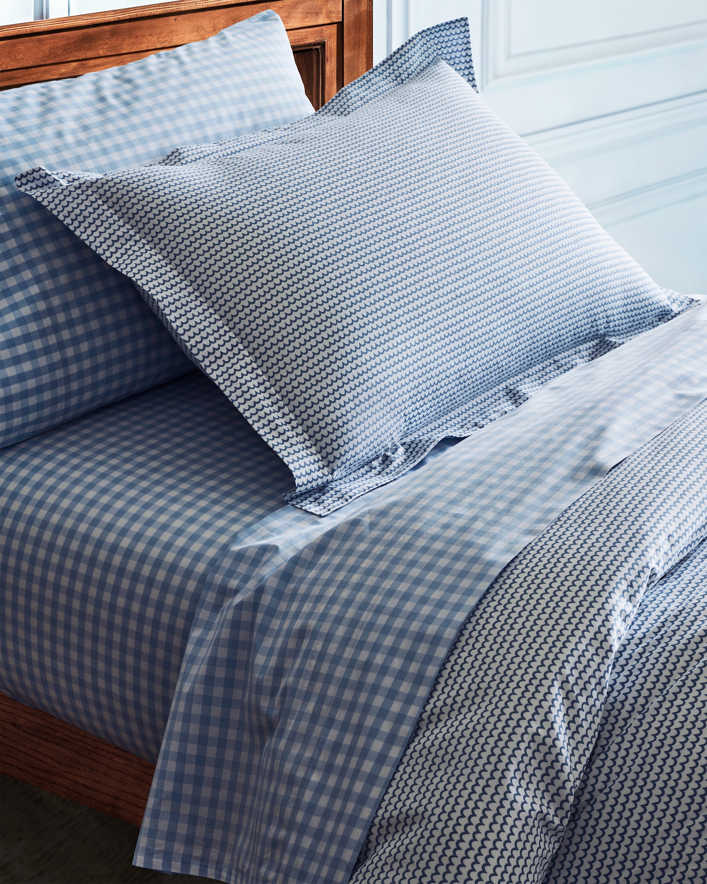 A neatly made bed with a wooden headboard features the Luxe Premium Percale Duvet Set in La Mer by Petite Plume, showcasing blue and white checks. A large geometric pillow sits on top. The 100% cotton set complements light-colored walls, creating a cozy atmosphere.