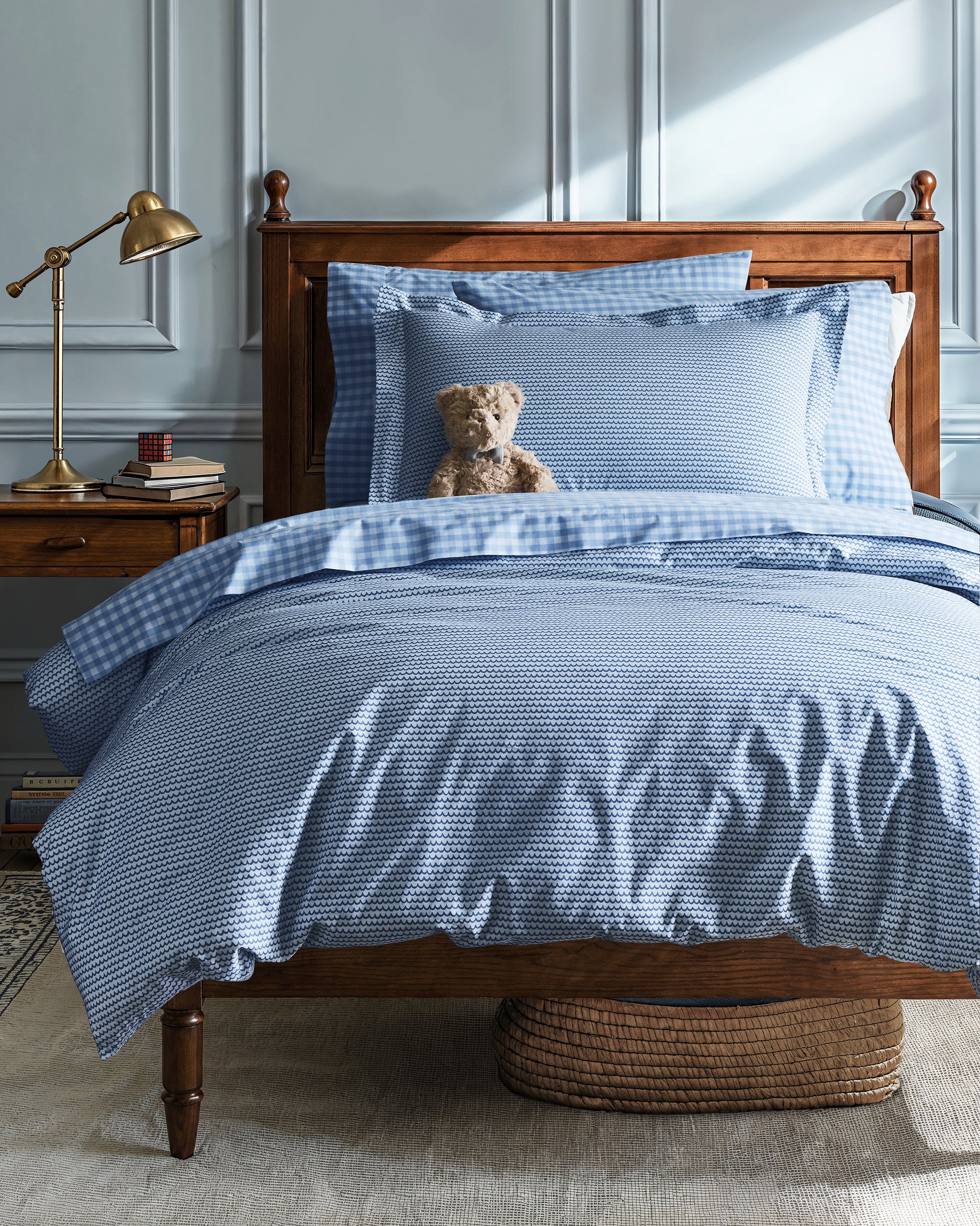 A cozy bedroom scene features a wooden bed adorned with the Luxe Premium Percale Duvet Set in La Mer by Petite Plume. A plush teddy bear leans on the pillows. Beside it, a wooden nightstand holds a brass lamp, books, and a clock as soft natural light streams through the window.