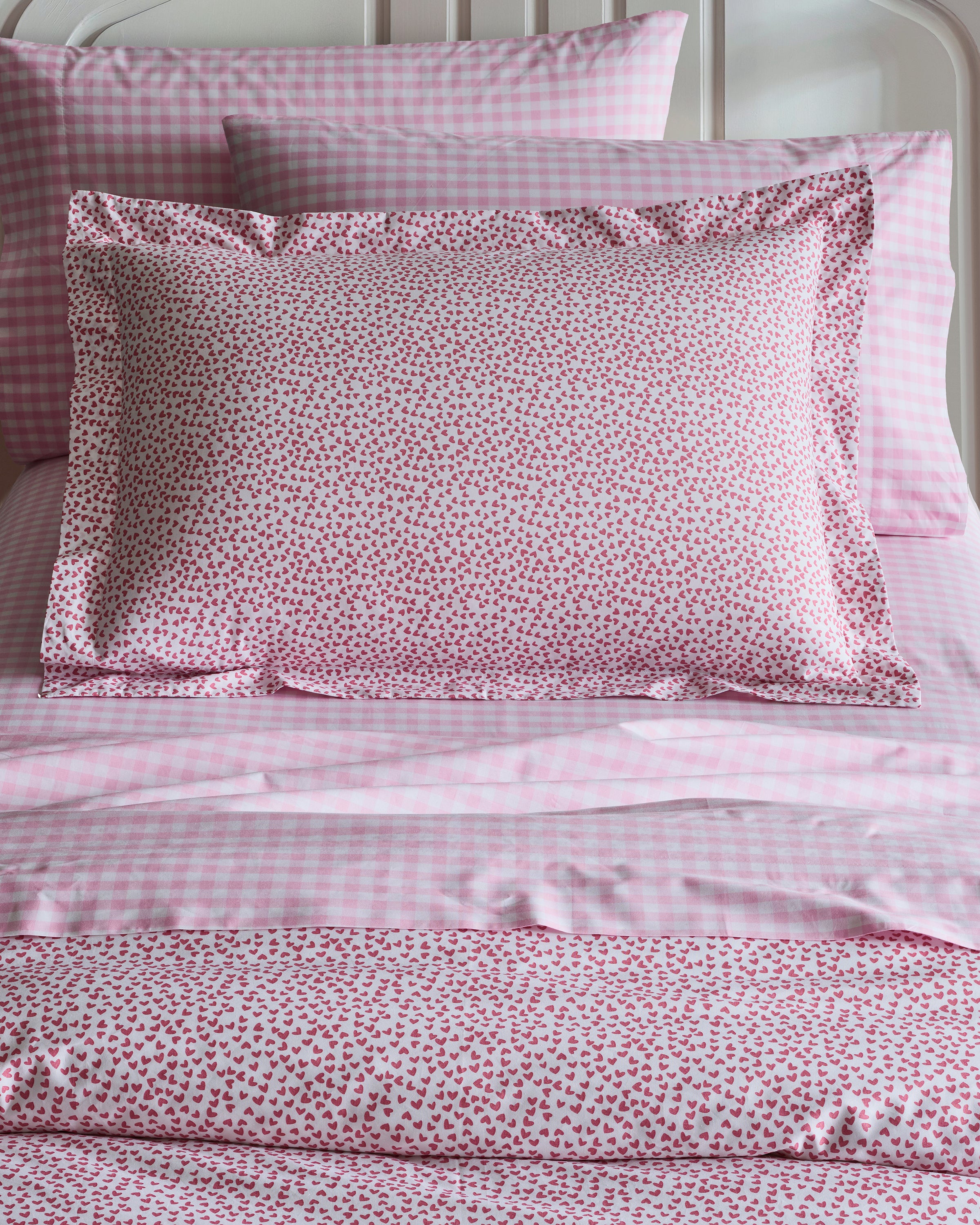 The Luxe Premium Percale Duvet Set in Sweethearts by Petite Plume features pink and white checkered 100% cotton sheets and pillowcases, topped with a heart-patterned decorative pillow. A white headboard and matching heart-themed accessories complete this luxurious look.