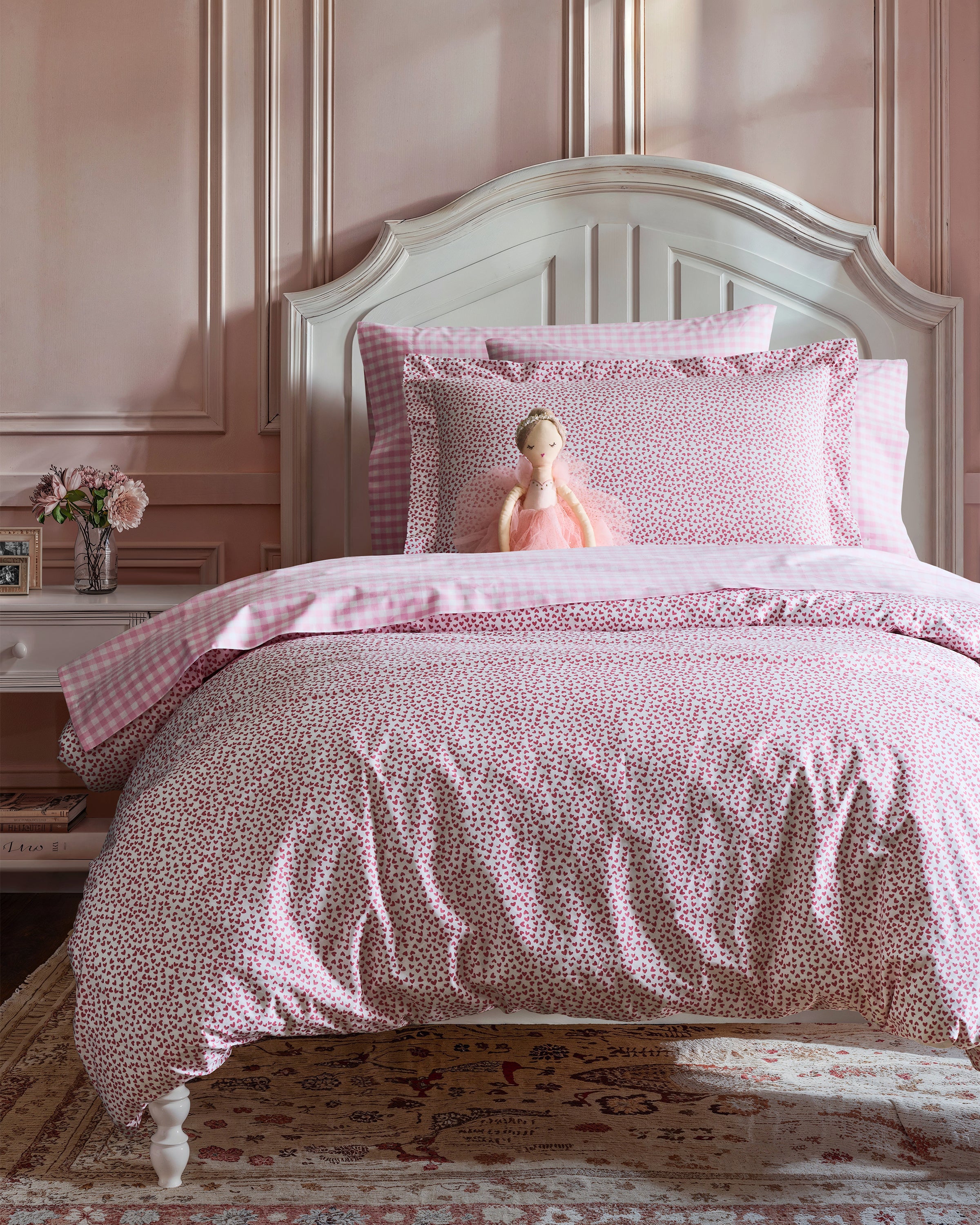 In a bedroom with a white bedframe and pink floral bedding, featuring the Luxe Premium Percale Duvet Set in Sweethearts by Petite Plume, a doll rests against the pillows. The wall has pink accents, while a bedside table holds pink flowers, and a patterned rug adorns the floor for an elegant touch.