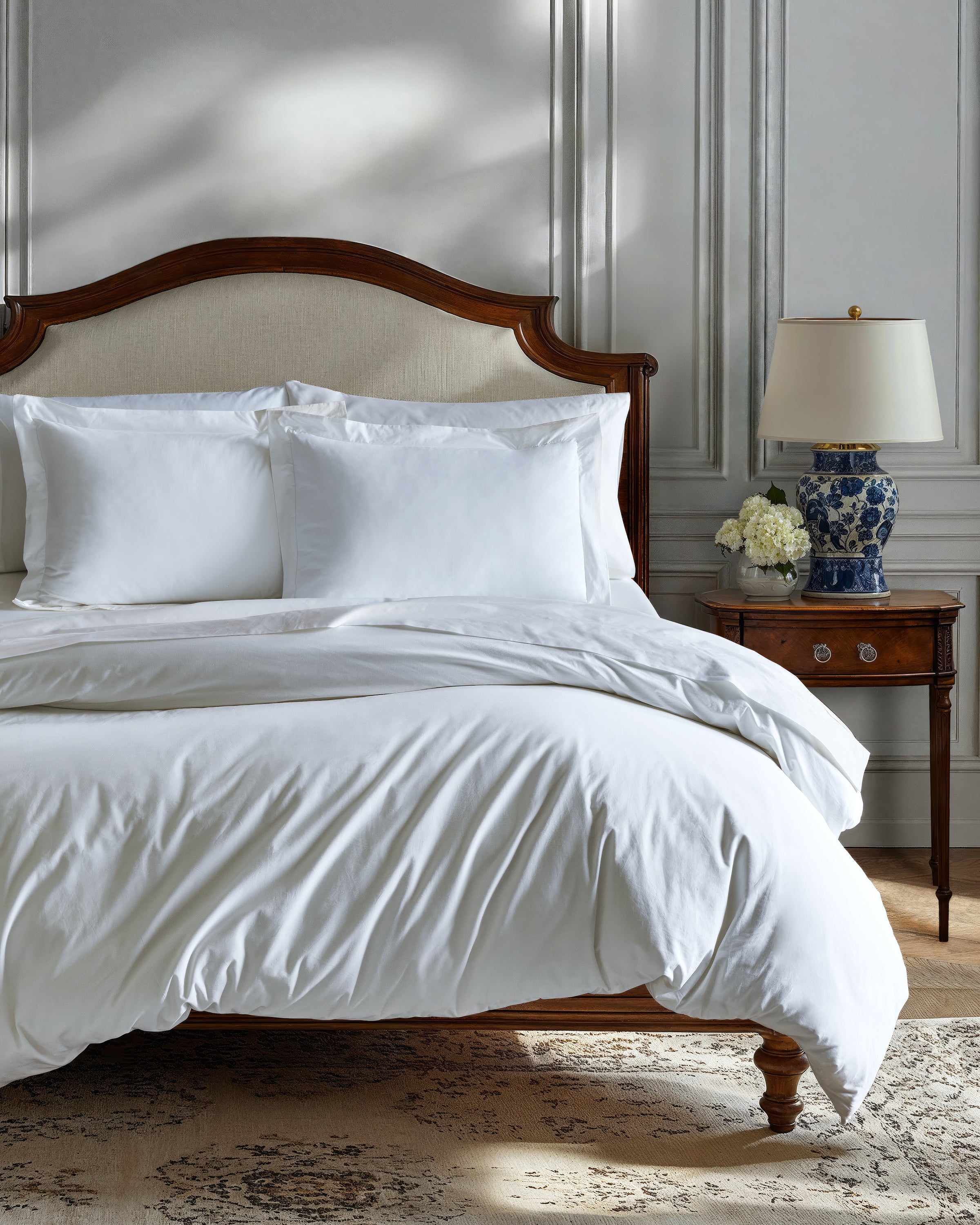 The elegant bedroom showcases a wooden bed with Petite Plumes Signature Luxe Sateen Bed Sheets in white and white pillows. A wooden side table features a blue-patterned lamp and floral arrangement, while classic paneled walls and a patterned rug enhance the five-star hotel suite ambiance.