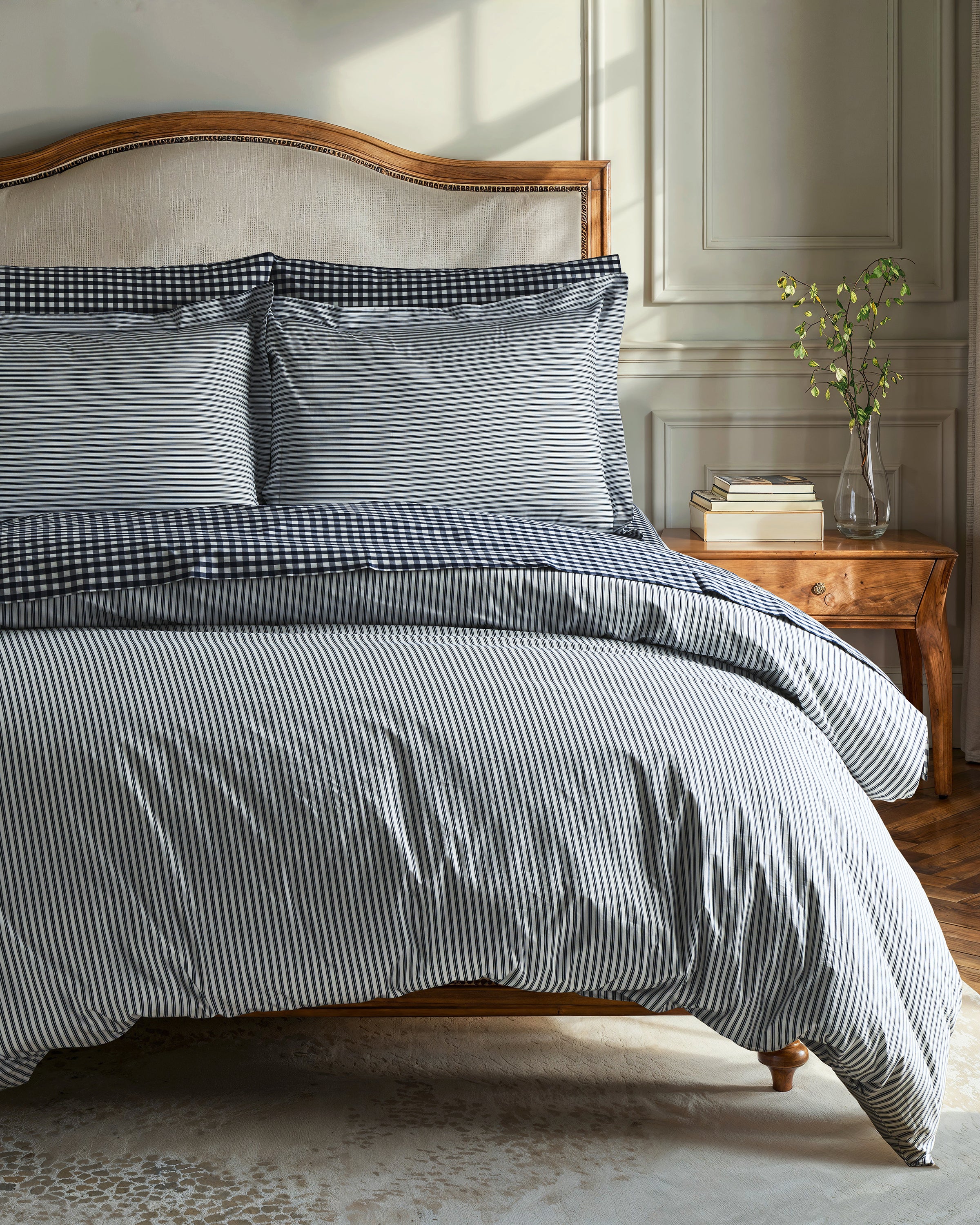 A neatly made bed with Petite Plumes Luxe Premium Percale Duvet Set in Navy French Ticking and two pillows stands against a light paneled wall. To the right, a wooden nightstand holds a vase with green branches, all bathed in soft natural light, highlighting the luxurious bedding.