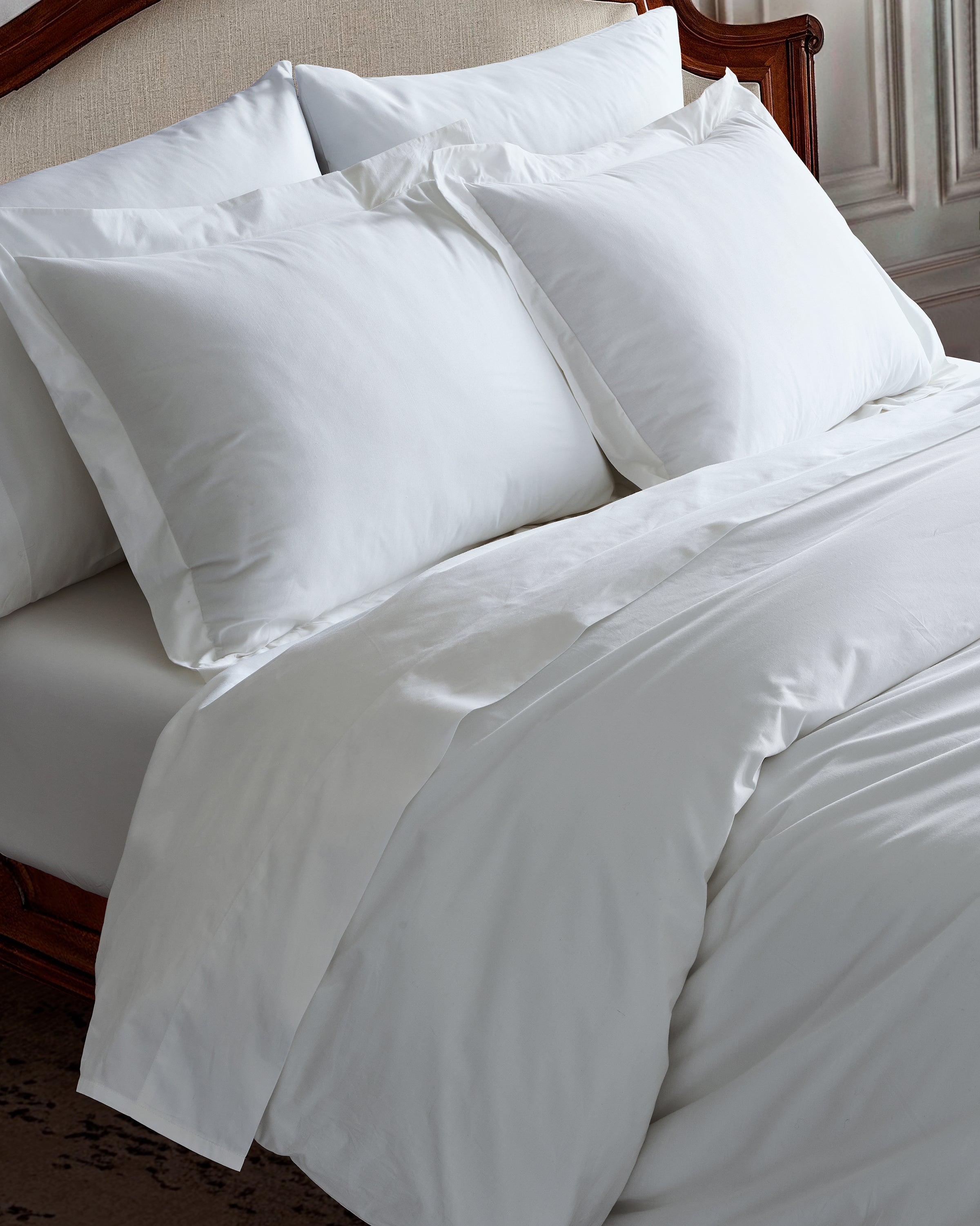 The bed features Petite Plume’s Signature Luxe Sateen Bed Sheets in White, complemented by a white comforter and pillows against a wooden headboard, creating an atmosphere akin to a softly lit, elegant five-star hotel room.