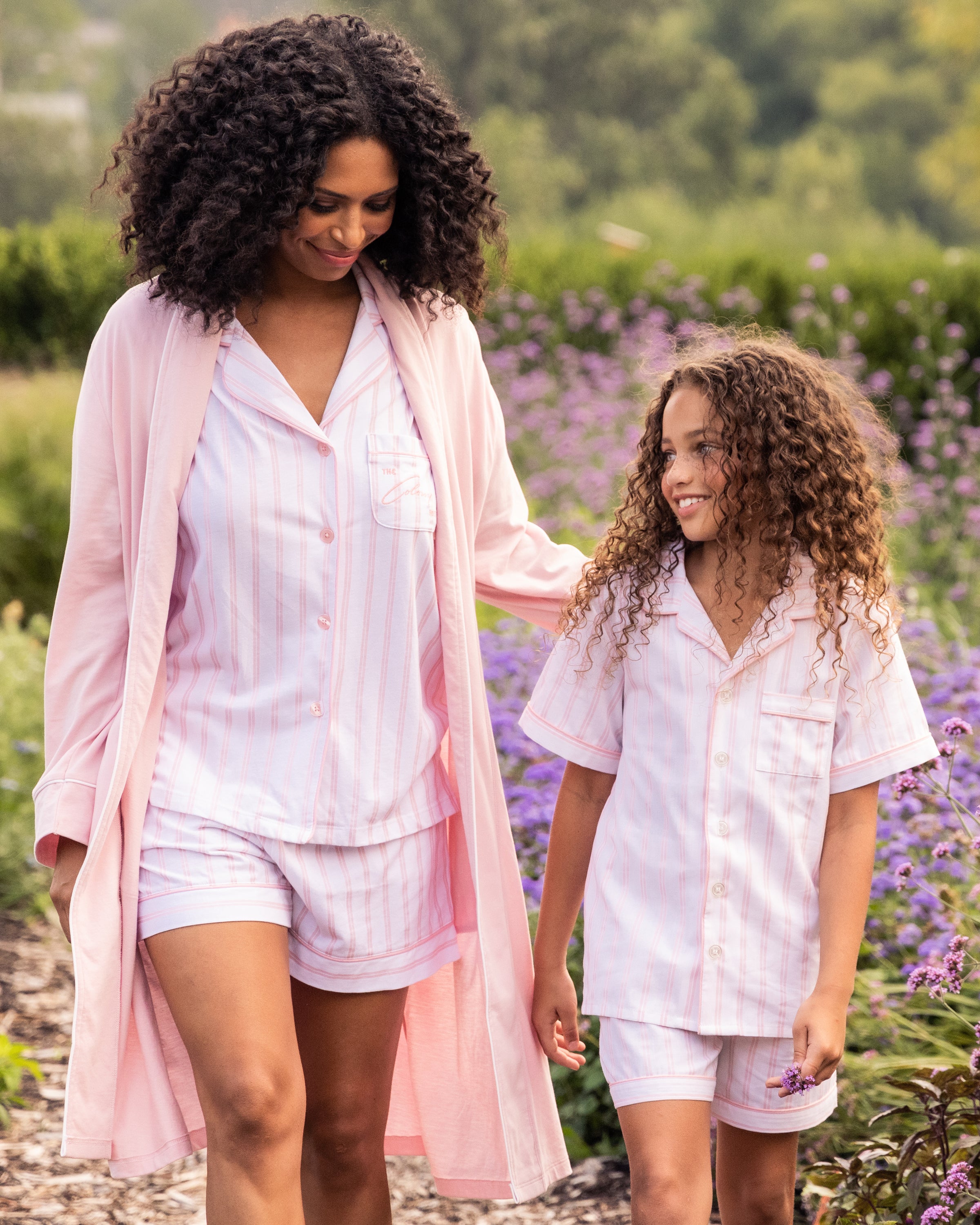 A woman and girl are smiling outdoors, dressed in matching Petite Plumes Kids Twill Pajama Short Set in Pink and White Stripe, made of luxurious cotton. The woman also wears a long robe amidst green foliage and purple flowers.