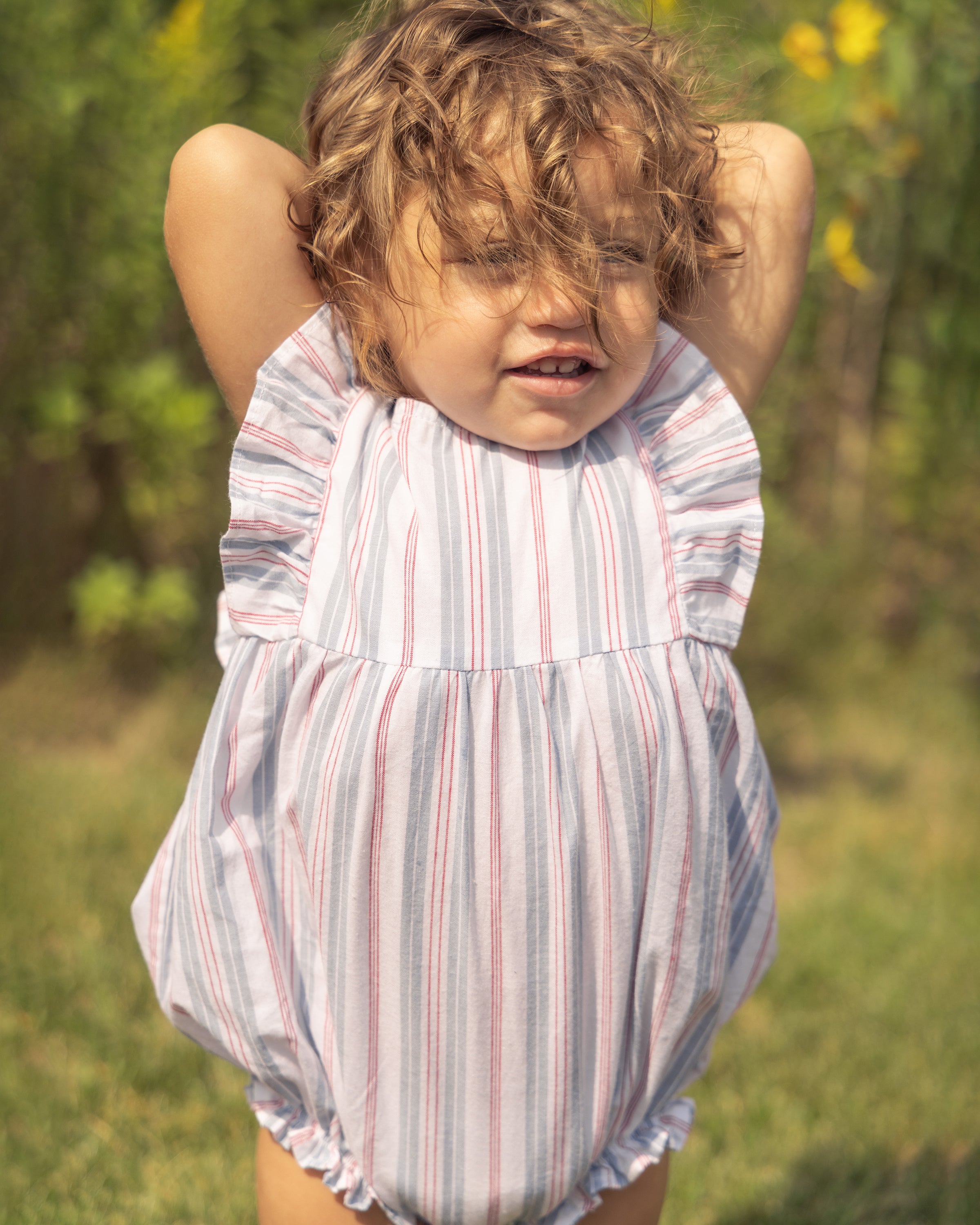 A young child with curly hair stands outside, wearing a Petite Plume Babys Twill Ruffled Romper in Vintage French Stripes. Their arms are raised as sunlight illuminates their face, casting shadows. The background features blurred green foliage and yellow flowers.