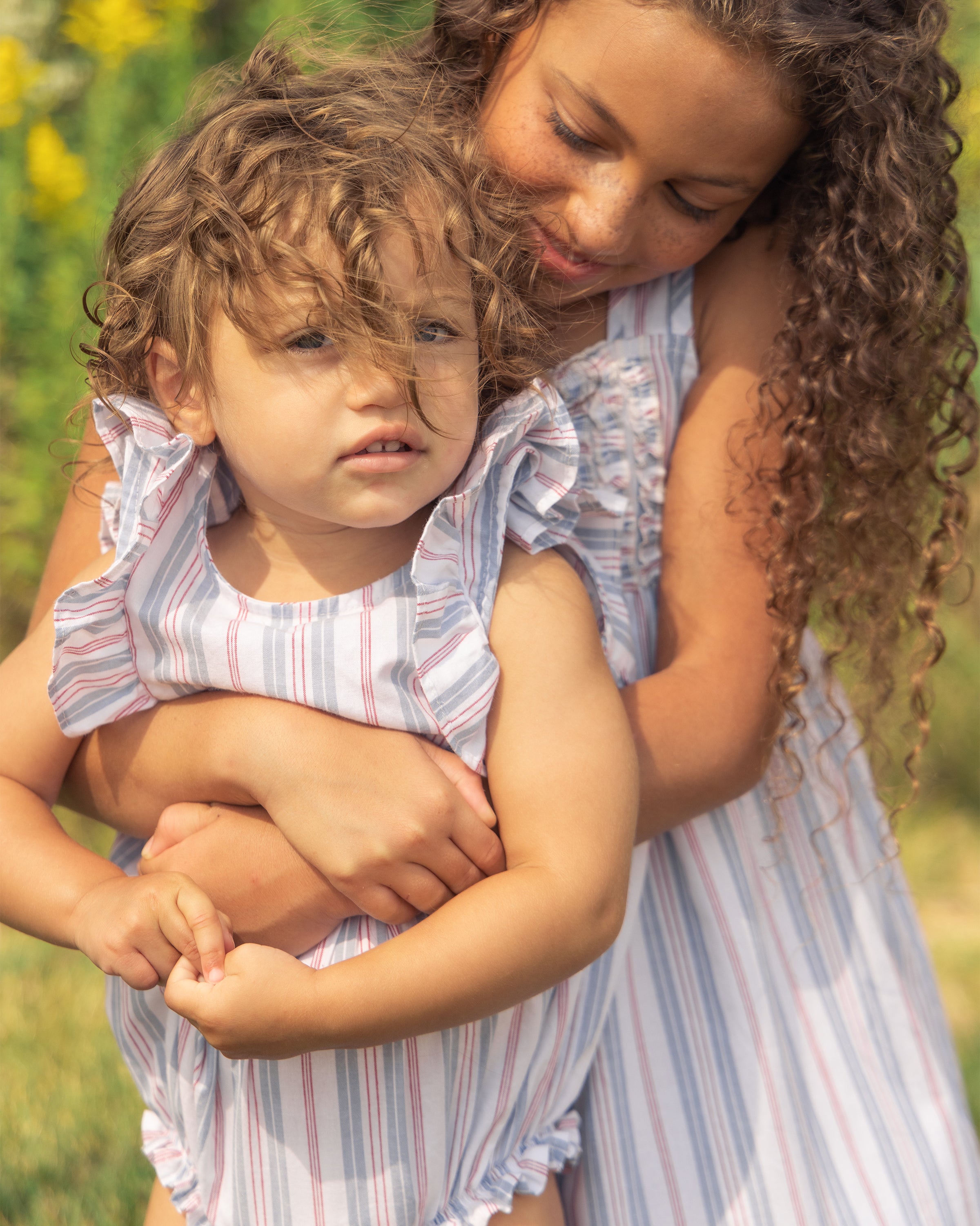 Two young children with curly hair stand in a sunny garden. The older child, wearing Petite Plumes Twill Ruffled Romper in Vintage French Stripes, hugs the younger from behind. Both wear pastel-colored dresses with a Victorian flair, against a backdrop of blurred green plants and yellow flowers.