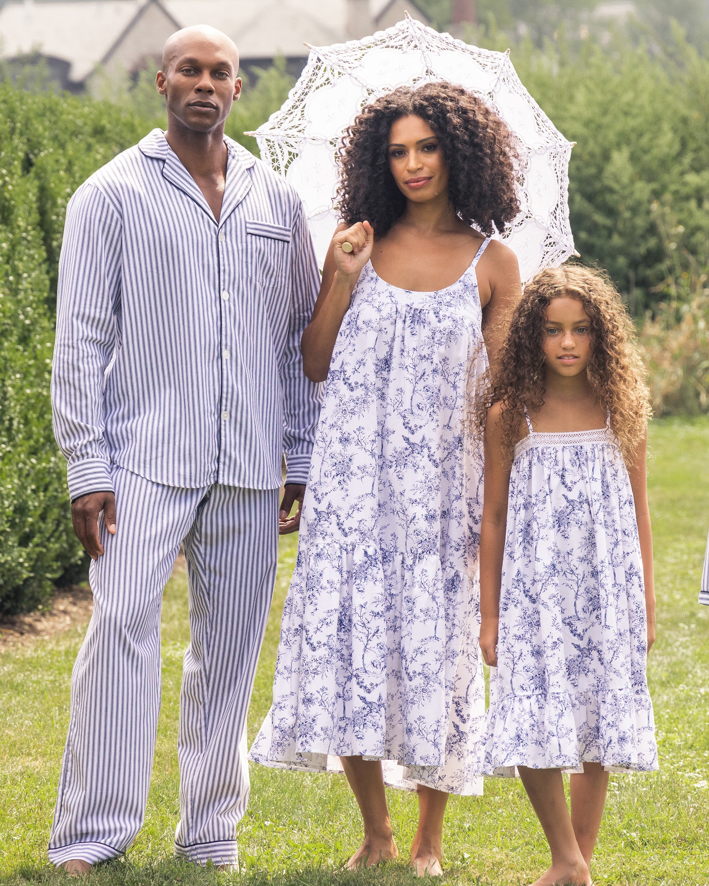 A family stands outdoors on grass. The man wears striped pajamas. The woman, holding a lace parasol, and the girl both wear matching floral dresses with a charming vintage print reminiscent of Petite Plumes Womens Twill Chloé Nightgown in Timeless Toile. Hedges and trees form a serene backdrop.