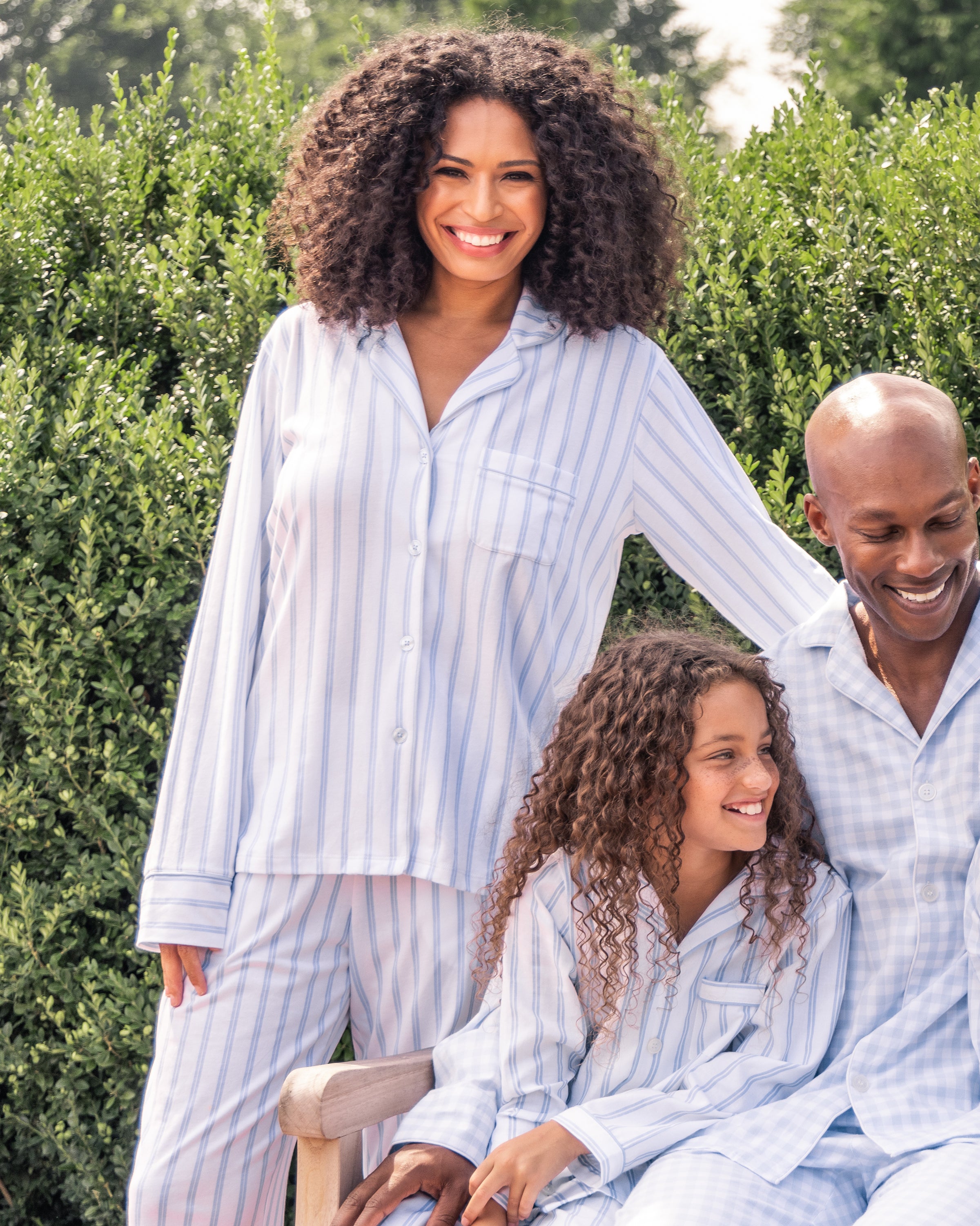 A woman, man, and child wear matching Petite Plume luxury pajamas in soft jersey knit. She smiles with her arm around him as he sits beside the child, who gazes up at him. Together, they share a joyful moment amidst lush green bushes.