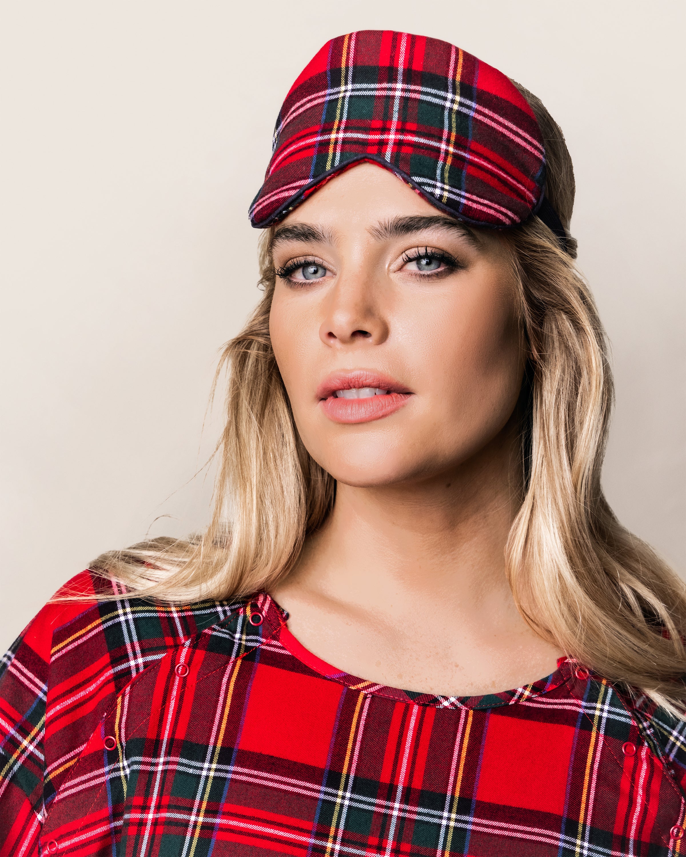 A woman with long blonde hair epitomizes luxury sleep accessories, wrapped in Petite Plumes red plaid sleep mask, the Adults Brushed Cotton Sleep Mask in Imperial Tartan, against a serene soft beige background.