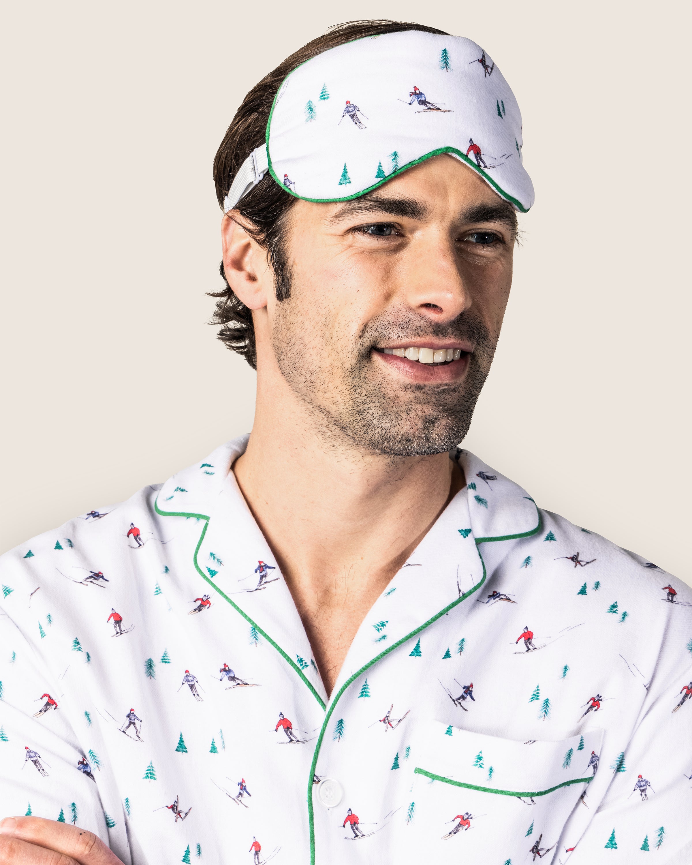 A man wears Petite Plumes white pajamas with green piping and an Adults Flannel Sleep Mask in Après Ski. Smiling brightly, he embodies luxury sleep. Add matching eye masks for a refined touch.