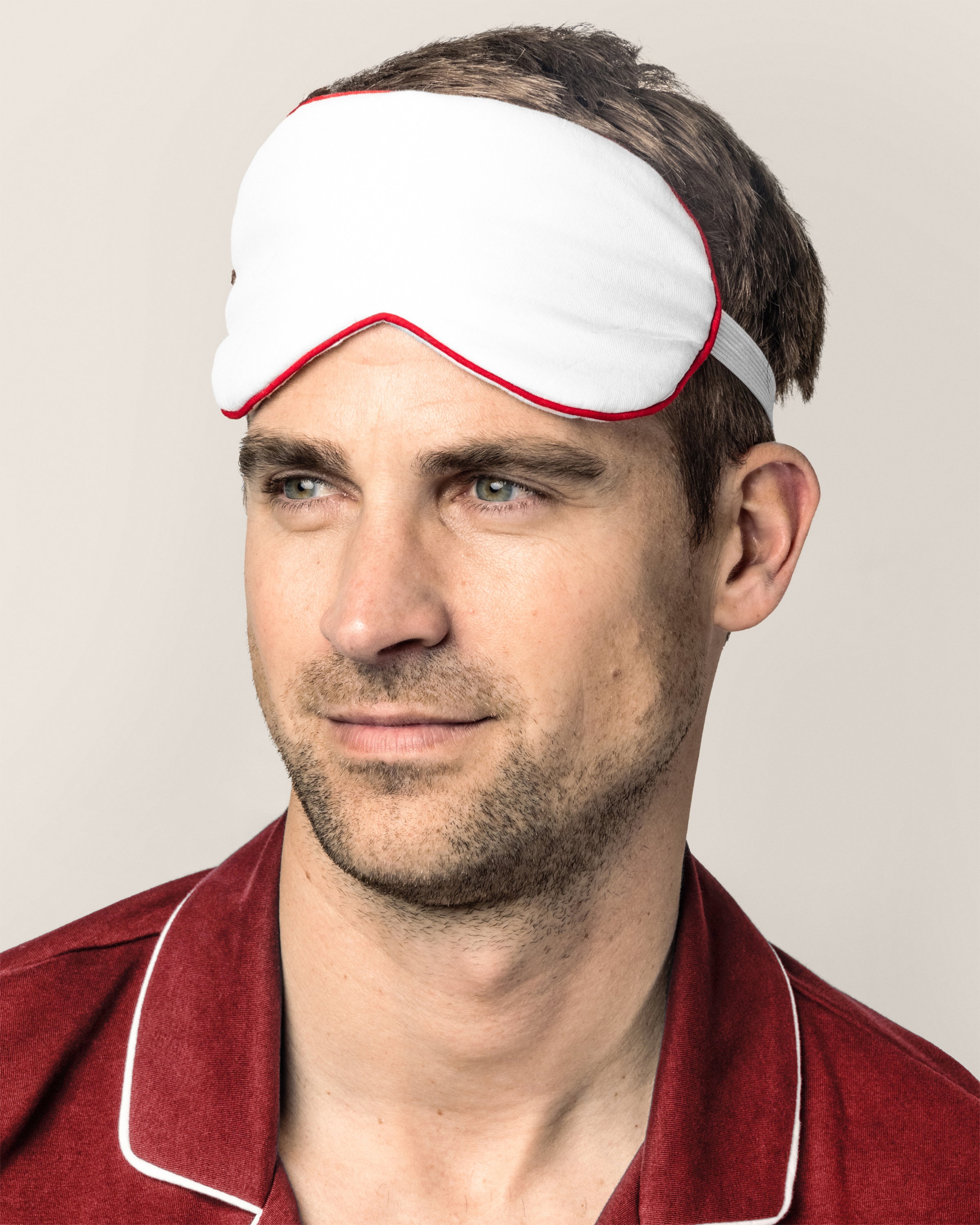 A man wearing Petite Plumes Adults Twill Sleep Mask in White with Red Piping gazes sideways. He pairs it with a matching red collared shirt against a light, plain background, highlighting his elegantly simple style.