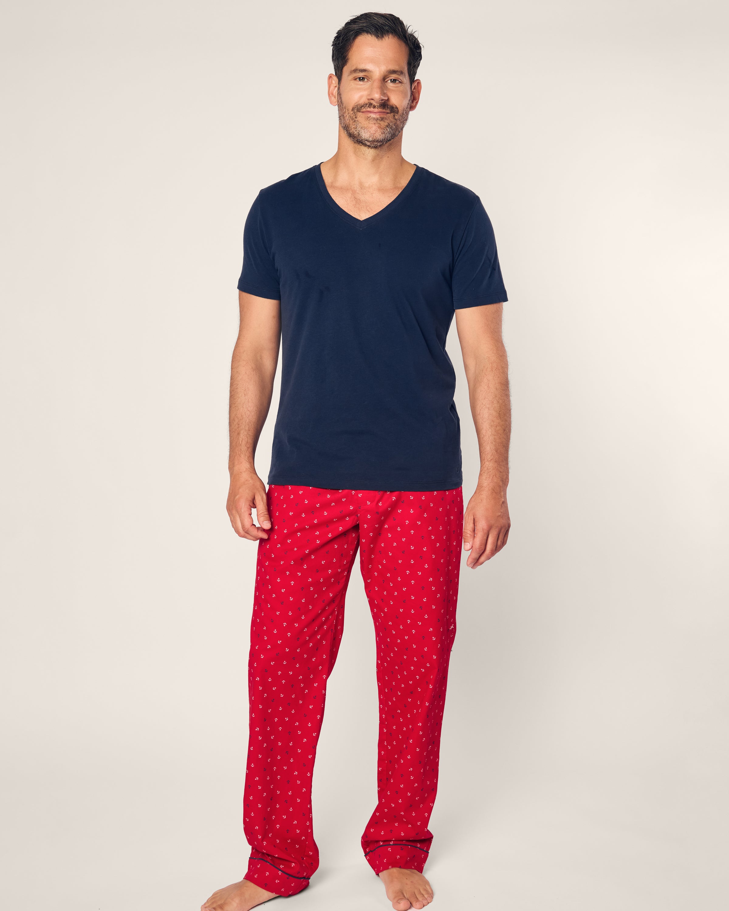 A smiling man stands barefoot on a plain background, wearing a black V-neck t-shirt and Petite Plumes Mens Twill Pajama Pant in Anchors Away, with his hands by his sides.