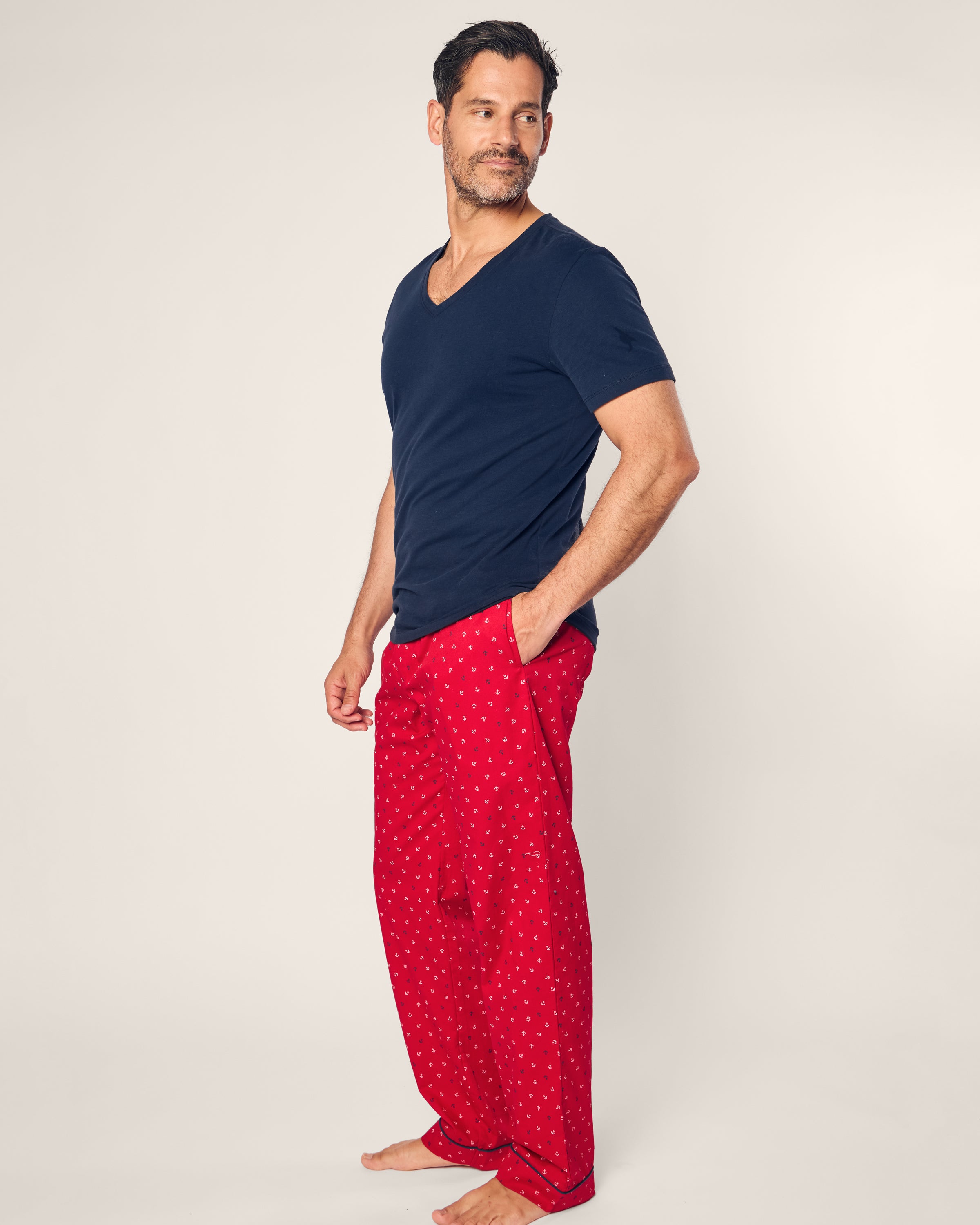 A man poses barefoot against a plain background, wearing a dark blue V-neck T-shirt and Petite Plumes Mens Twill Pajama Pant in Anchors Away, crafted from premium cotton with a small white print. With one hand in his pocket, he gazes thoughtfully to the side.