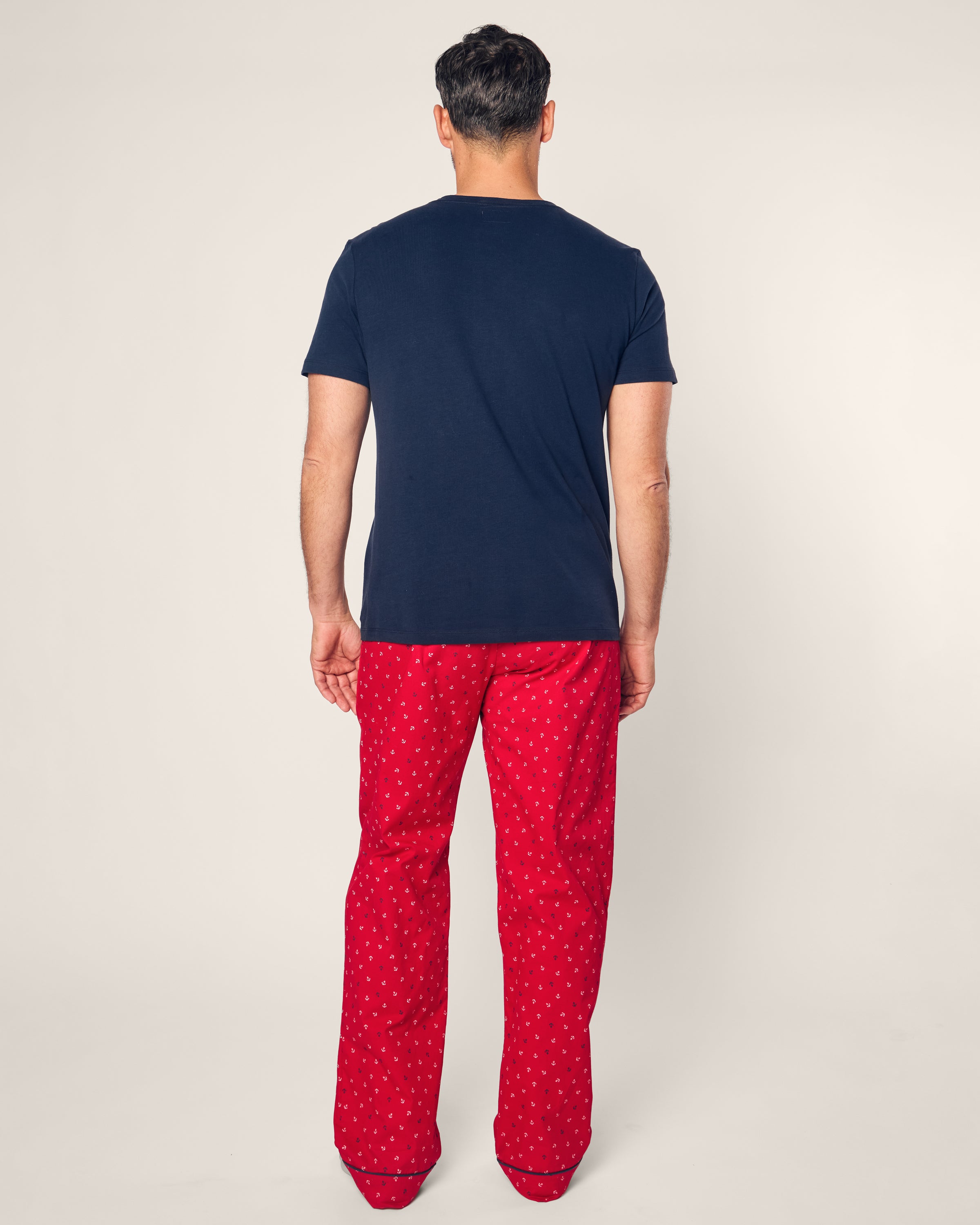 A man stands facing away, wearing a dark blue T-shirt and Petite Plumes Mens Twill Pajama Pant in Anchors Away print. The pants are red with white polka dots, crafted from high-quality yarn-dyed cotton against a plain light background.