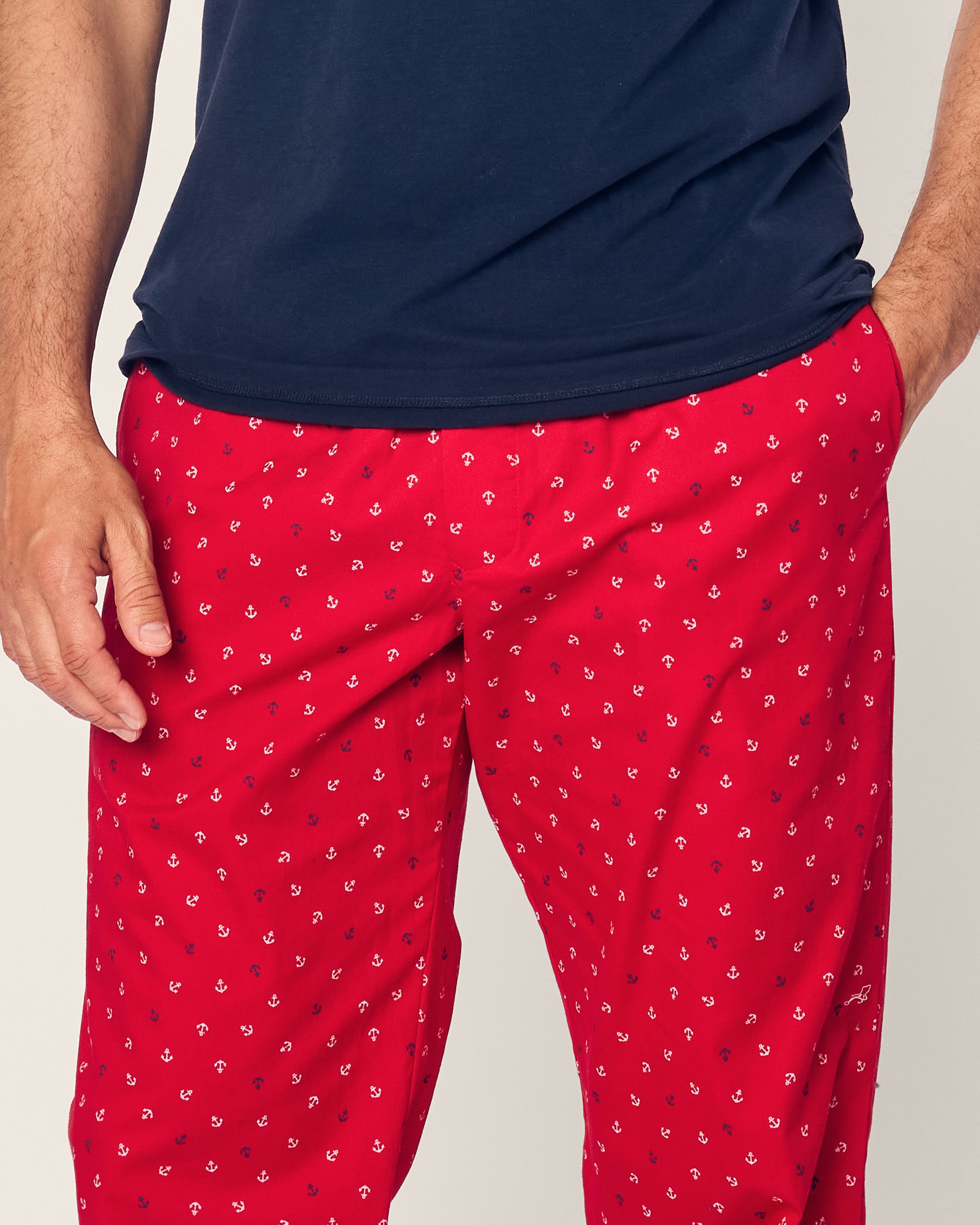 A person dressed in Petite Plumes Mens Twill Pajama Pant in Anchors Away, featuring the subtle print on premium navy blue fabric, is seen with a hand casually in their pocket. Only the lower part of the shirt and red pants are visible, showcasing their yarn-dyed richness.