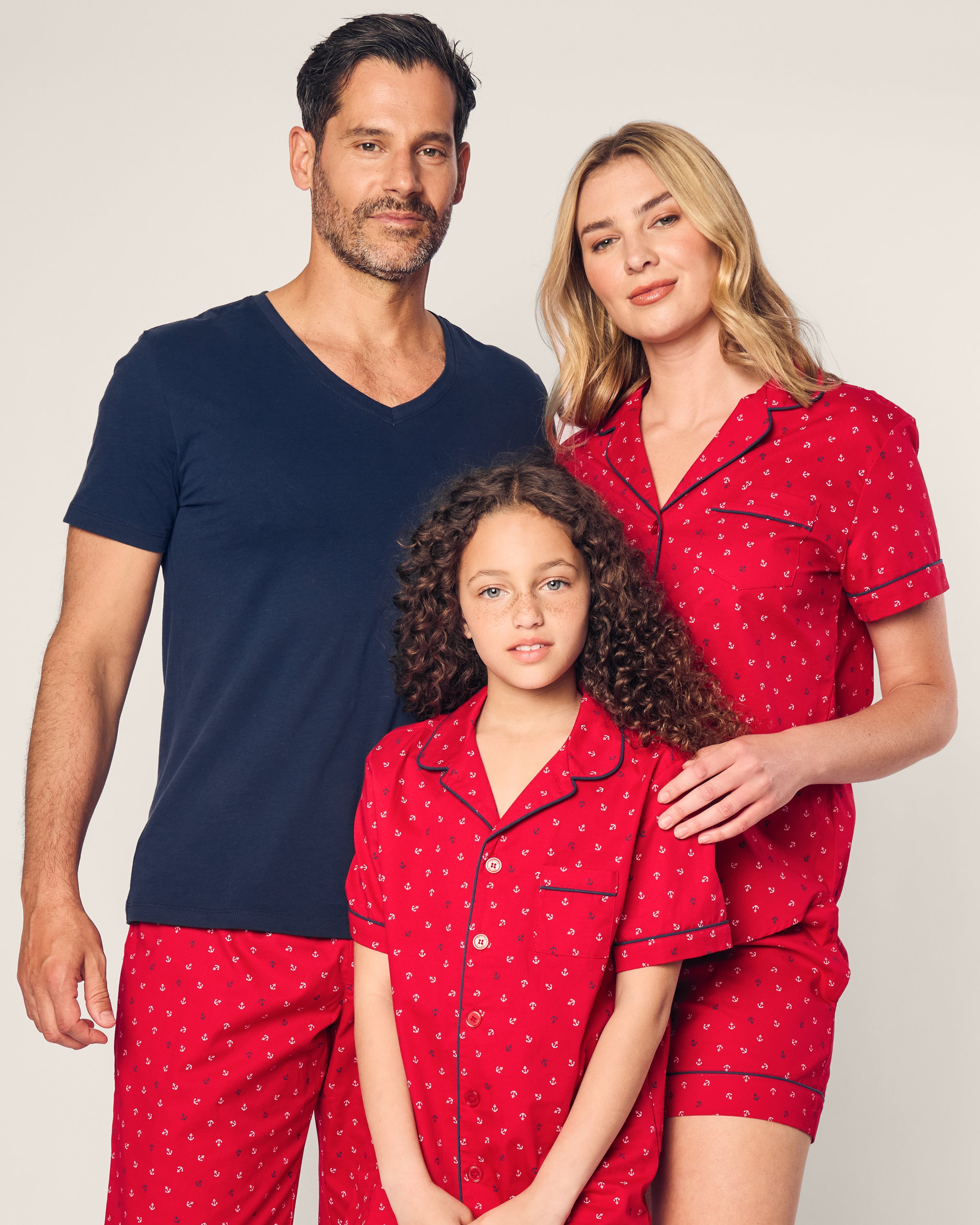 A man in a navy T-shirt and red shorts joins a woman and girl in matching Petite Plumes finest cotton pajamas with Anchors Away print. The woman has long blonde hair, the girl has curly hair, and they all smile warmly against a neutral background.