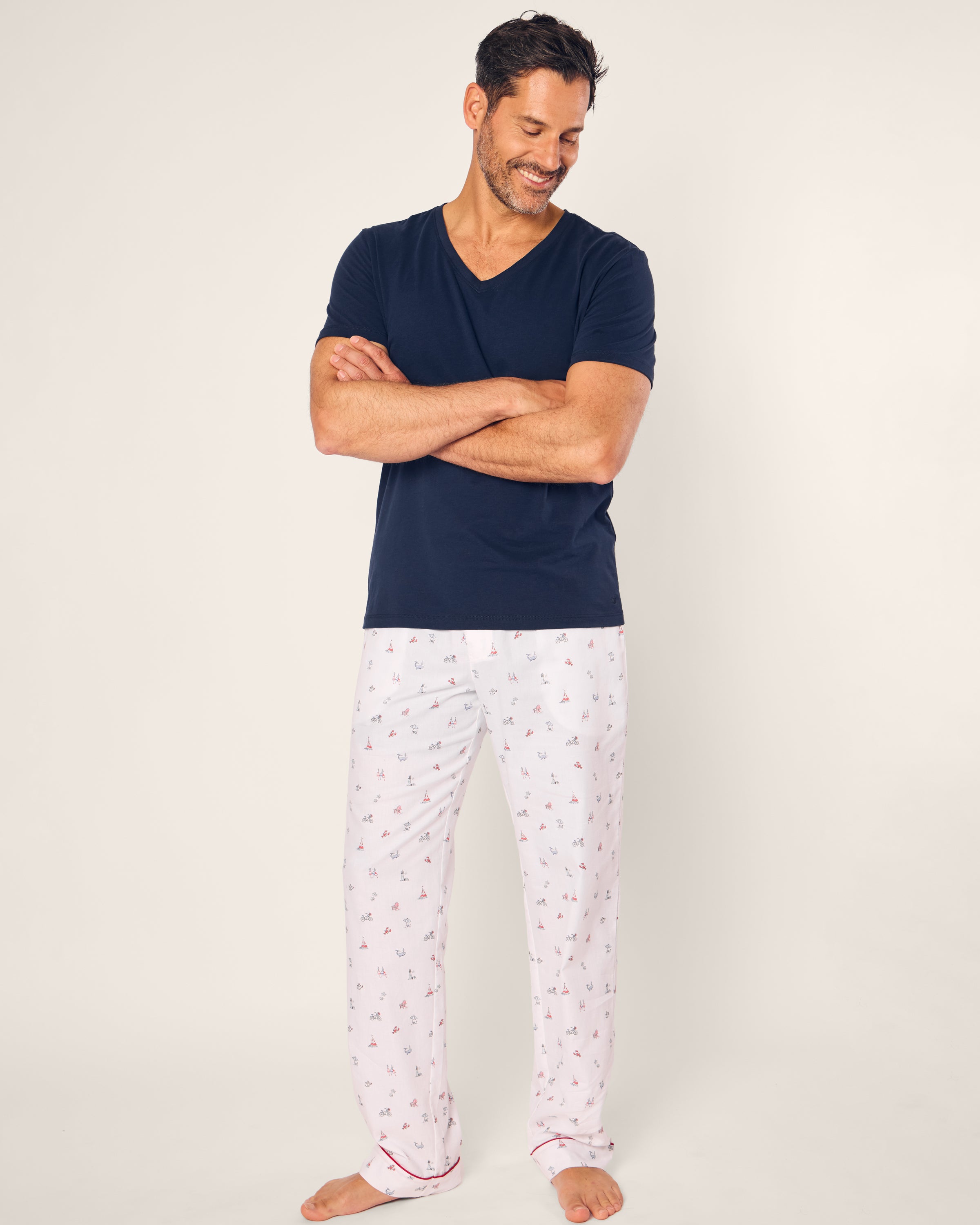 A person stands smiling with arms crossed, wearing a navy blue V-neck T-shirt and Petite Plumes Mens Twill Pajama Pant in Coastal Charms. The cotton sleepwear exudes comfort against the plain light gray background.