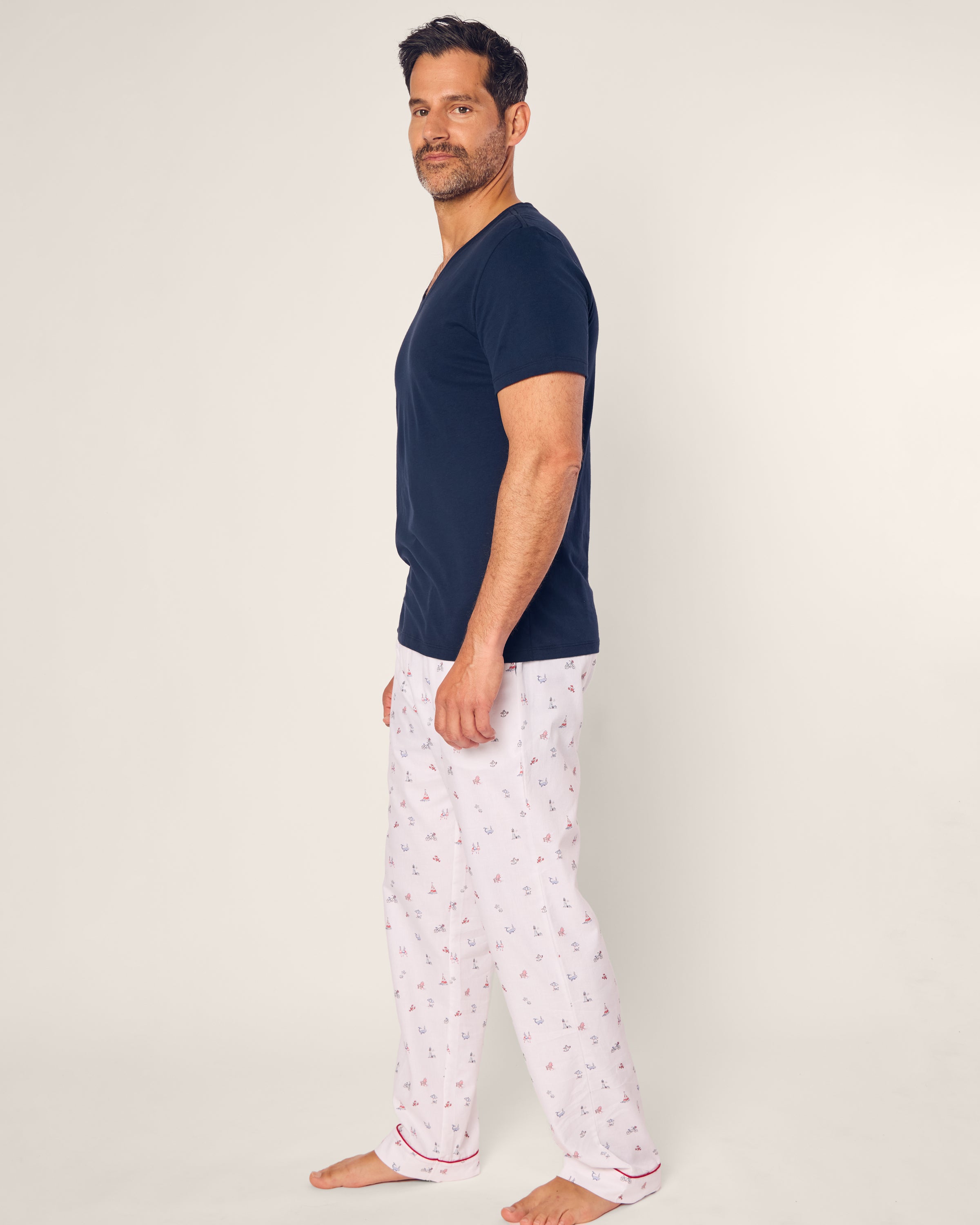 A man in a navy T-shirt and Petite Plumes Coastal Charms twill pajama pants stands barefoot against a plain background, embodying the ease of coastal charms as he looks at the camera.