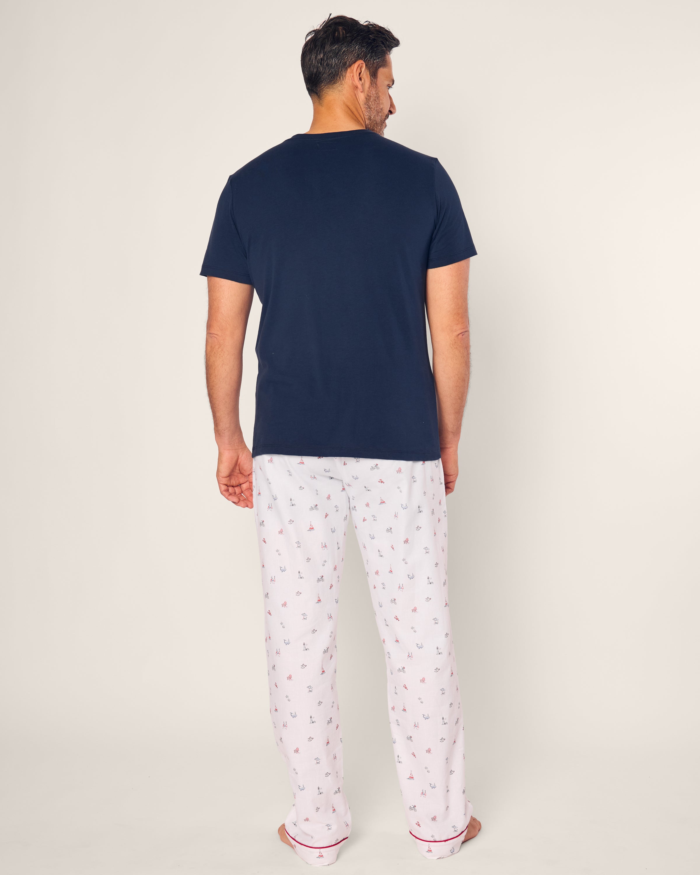 A person stands with their back to the camera, wearing a Petite Plume dark navy T-shirt and Mens Twill Pajama Pant in Coastal Charms. The cotton sleepwear suggests comfort and style against the plain, light-colored background.