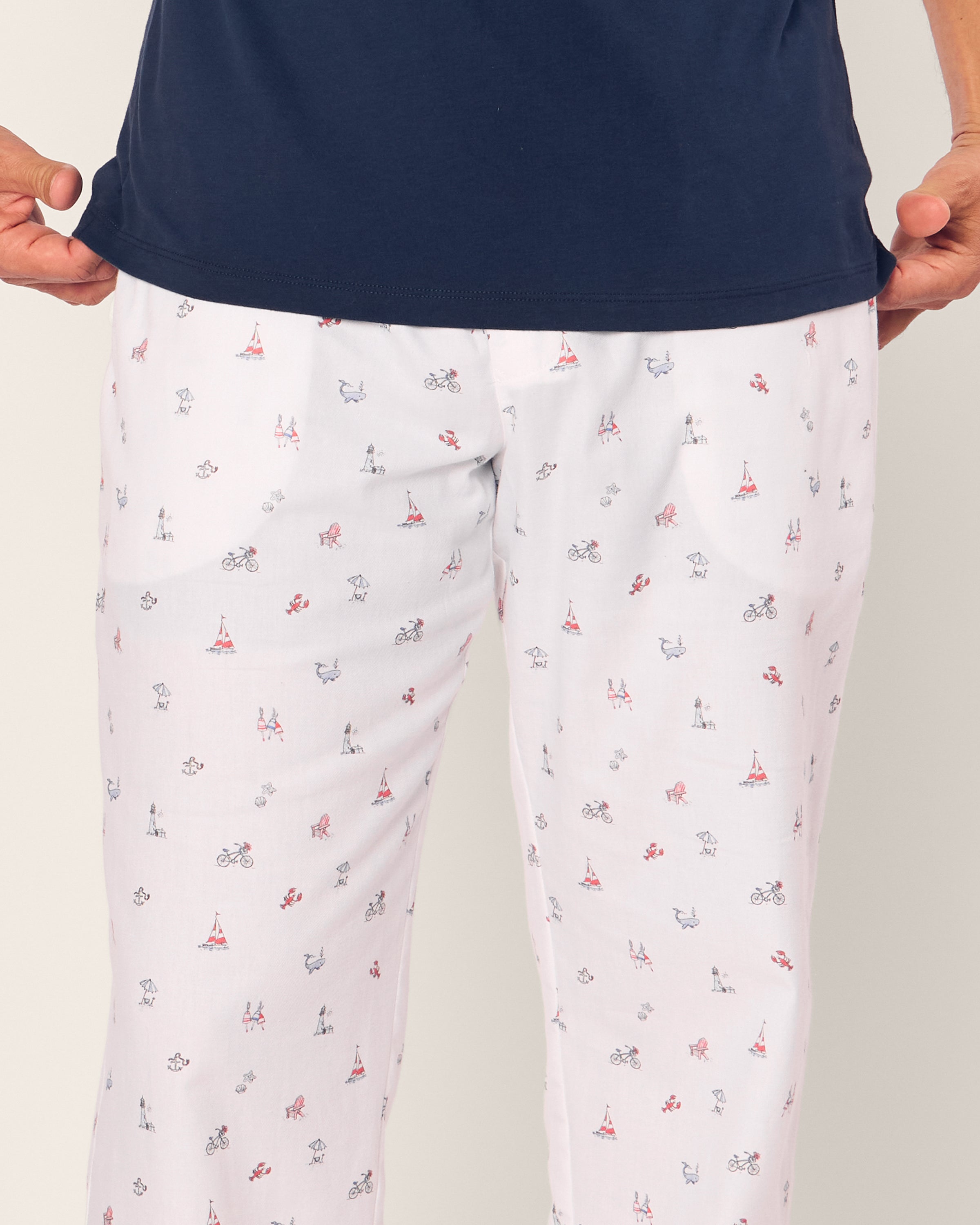 A person in luxurious cotton sleepwear wears a Petite Plume dark blue top and Mens Twill Pajama Pants in Coastal Charms, featuring playful prints of bicycles, Eiffel Towers, poodles, and red shoes.