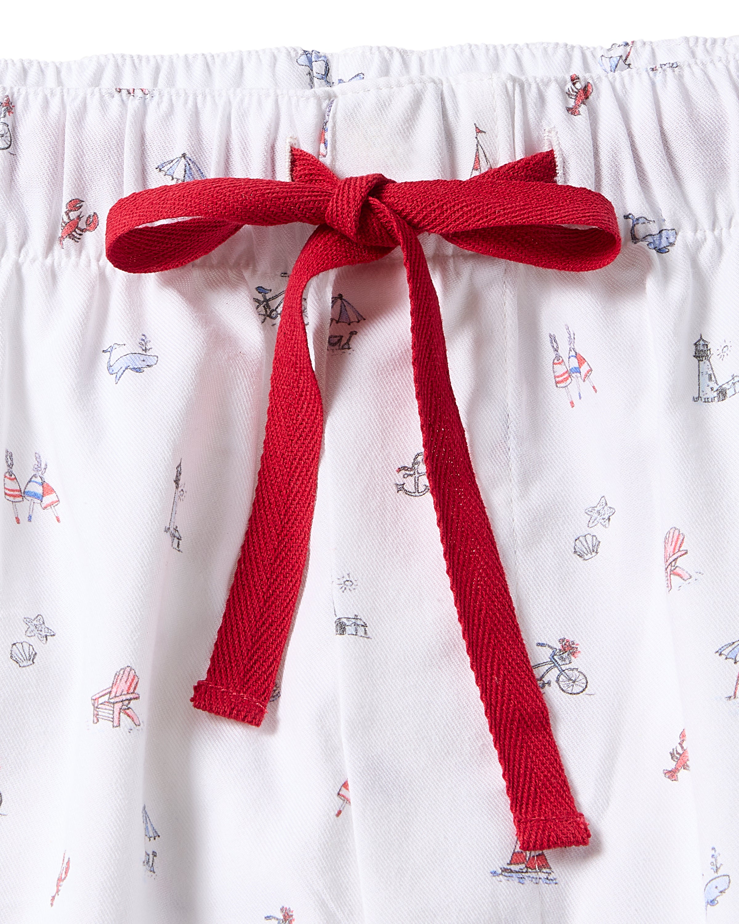 The Petite Plume Mens Twill Pajama Pant in Coastal Charms features white cotton sleepwear with small red and blue nautical-themed illustrations, including boats, chairs, and lighthouses. A central red decorative bow adds a striking contrast to the design.