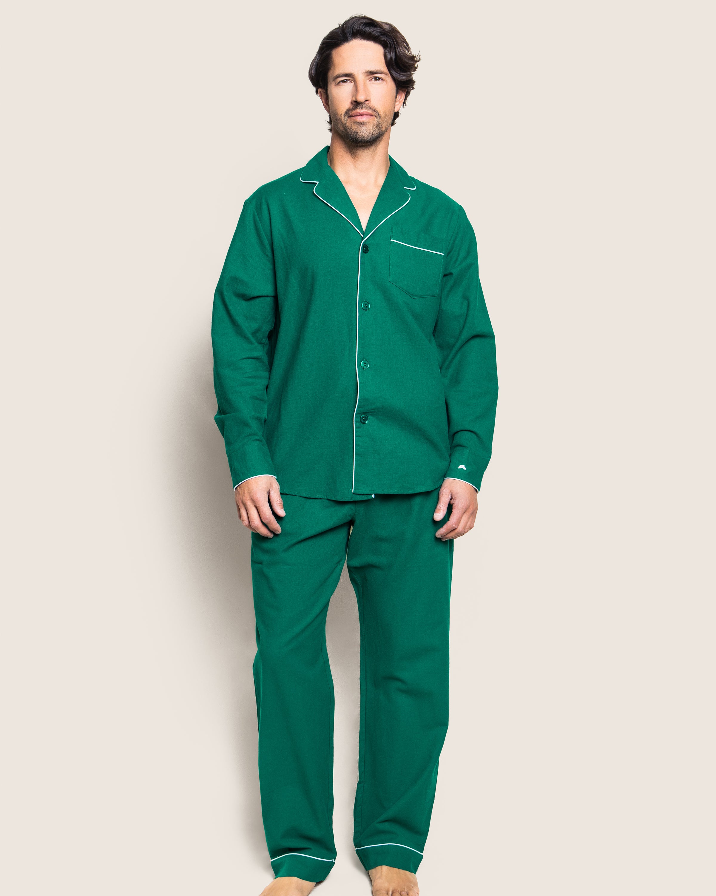 A man stands against a plain background wearing Petite Plumes Mens Flannel Pajama Set in Forest Green, featuring white piping. He has short, dark hair and a beard. The top includes a breast pocket and buttons down the front, while the pants are loose-fitting.