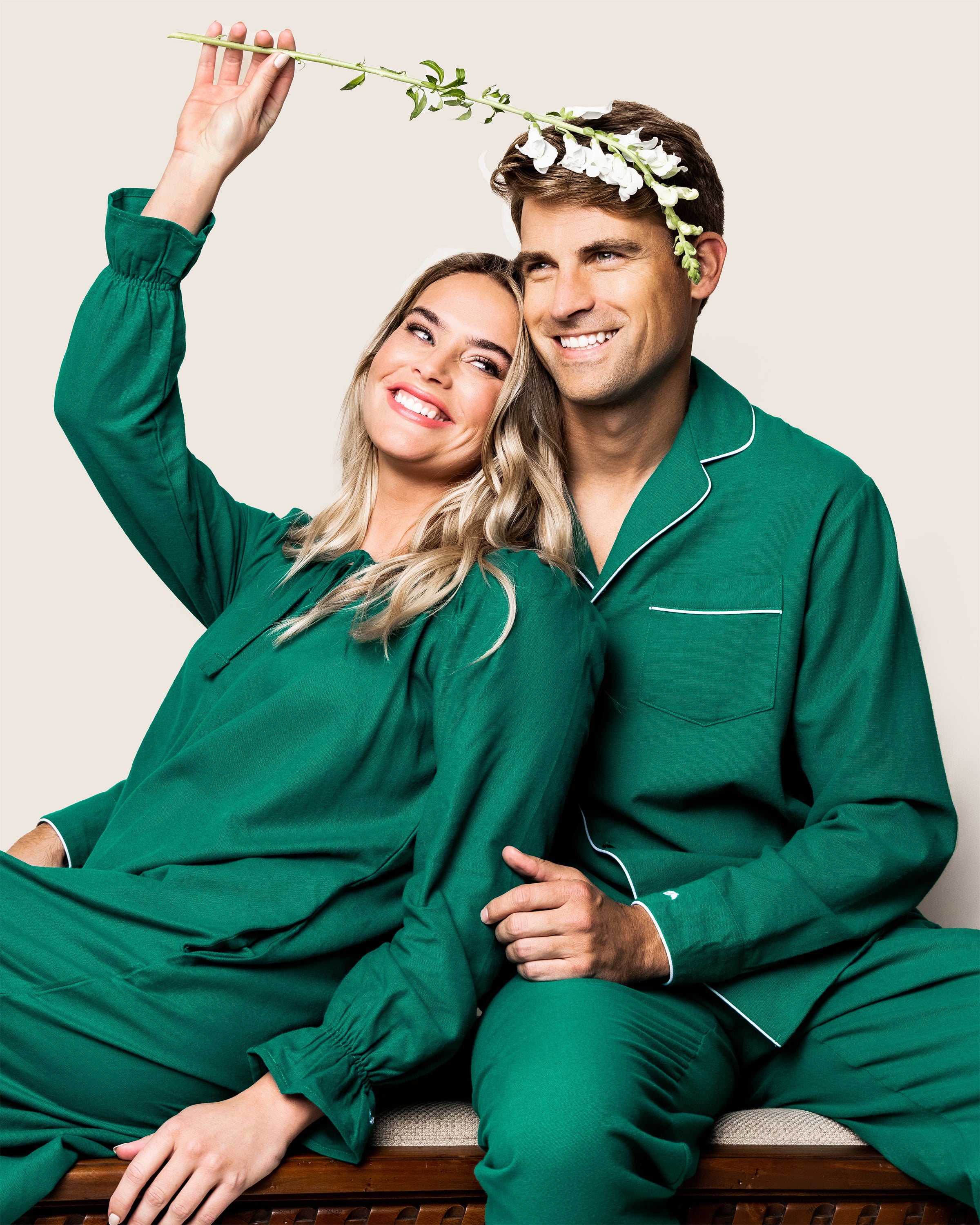 A woman in a green cotton pajama set playfully holds a flower over the head of a man dressed in Petite Plumes Mens Flannel Pajama Set in Forest Green. They both smile while seated against a simple backdrop.