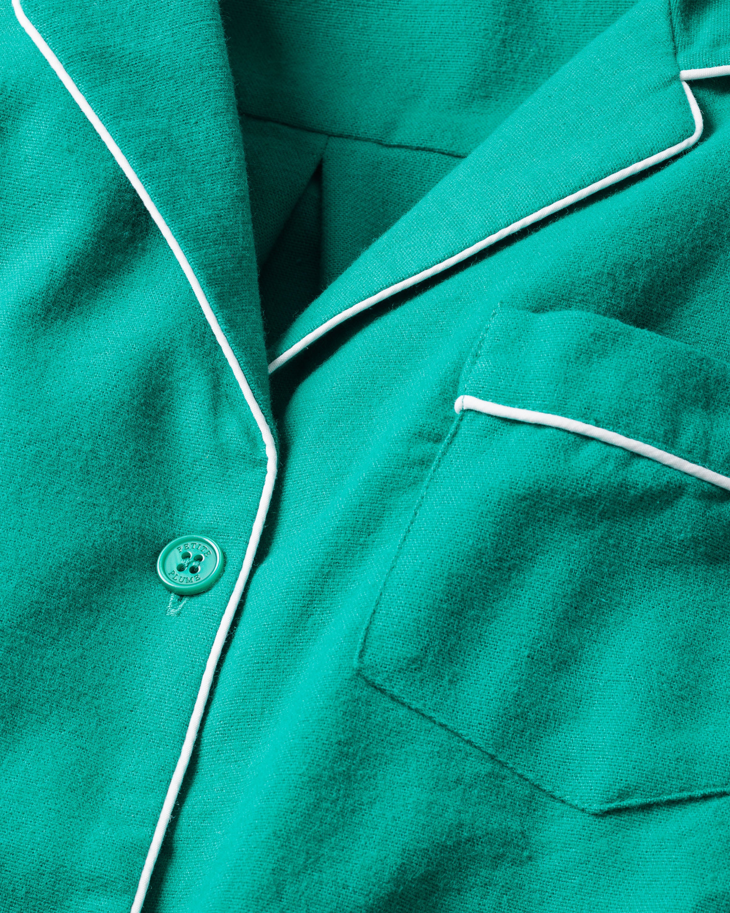 Close-up of a luxurious Petite Plume piece: the Mens Flannel Pajama Set in Forest Green, featuring white piping. The image highlights a single button, part of the collar, and a left-side pocket. Made from 100% cotton, the fabric appears soft and comfortable.