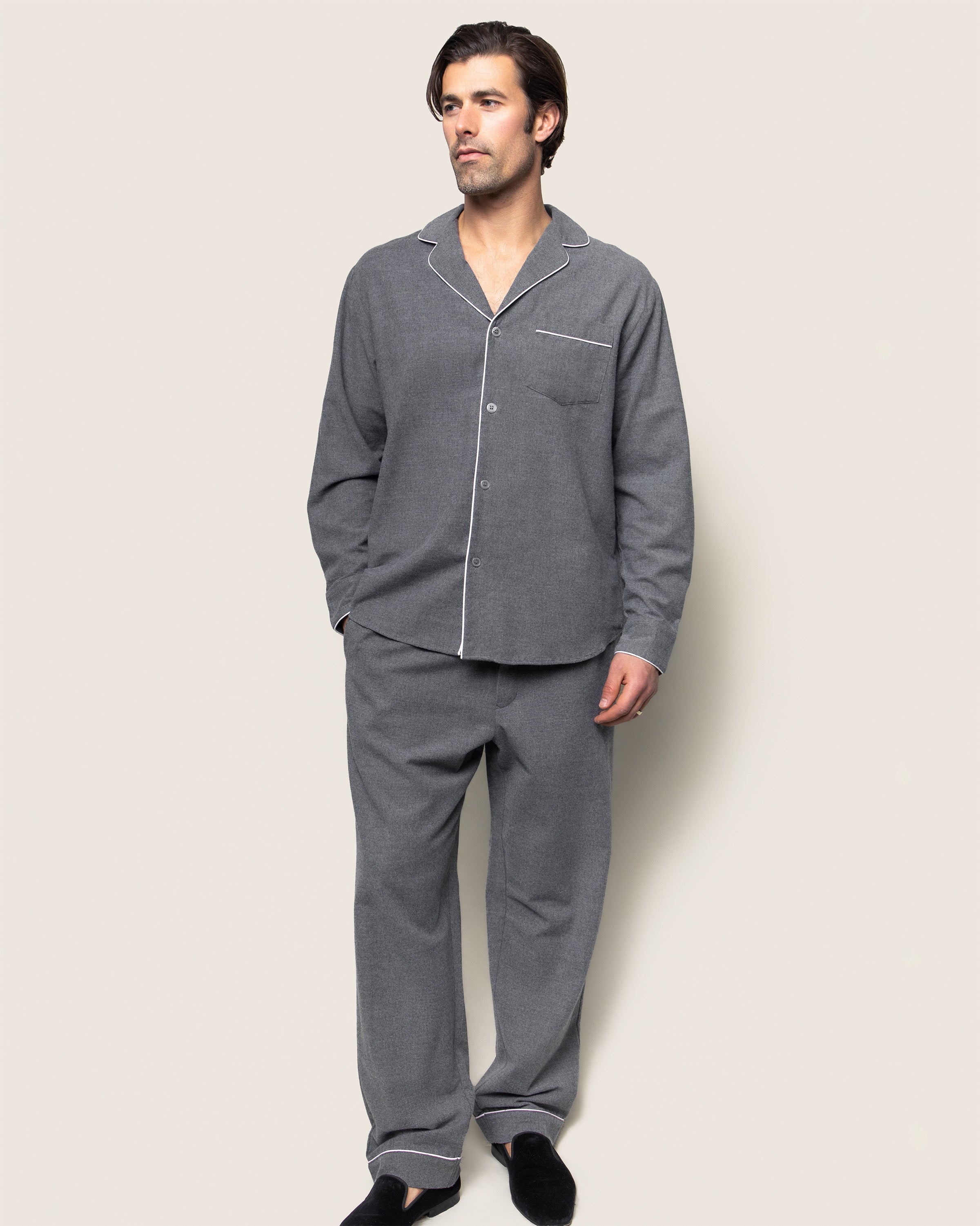 A man with short dark hair stands against a plain background, wearing Petite Plumes Mens Flannel Pajama Set in Grey. The cozy cotton twill set with white piping includes a long-sleeve top with collar and front pocket and matching pants, complemented by black slippers for stylish comfort.