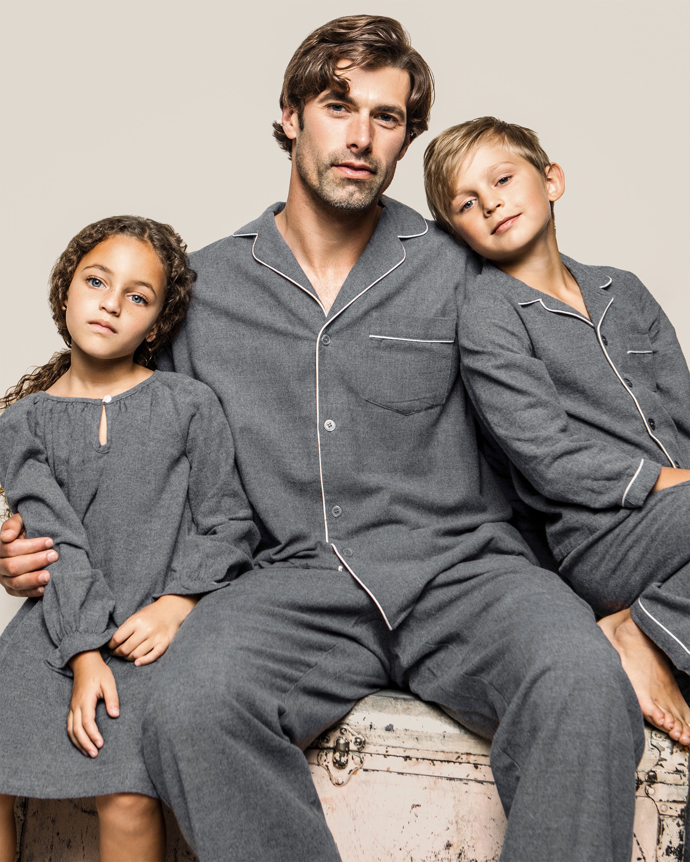 A man in a grey Mens Flannel Pajama Set by Petite Plume hugs two children. The girl wears a grey dress, and the boy matches in pajamas. Theyre seated on a vintage trunk against a plain background.