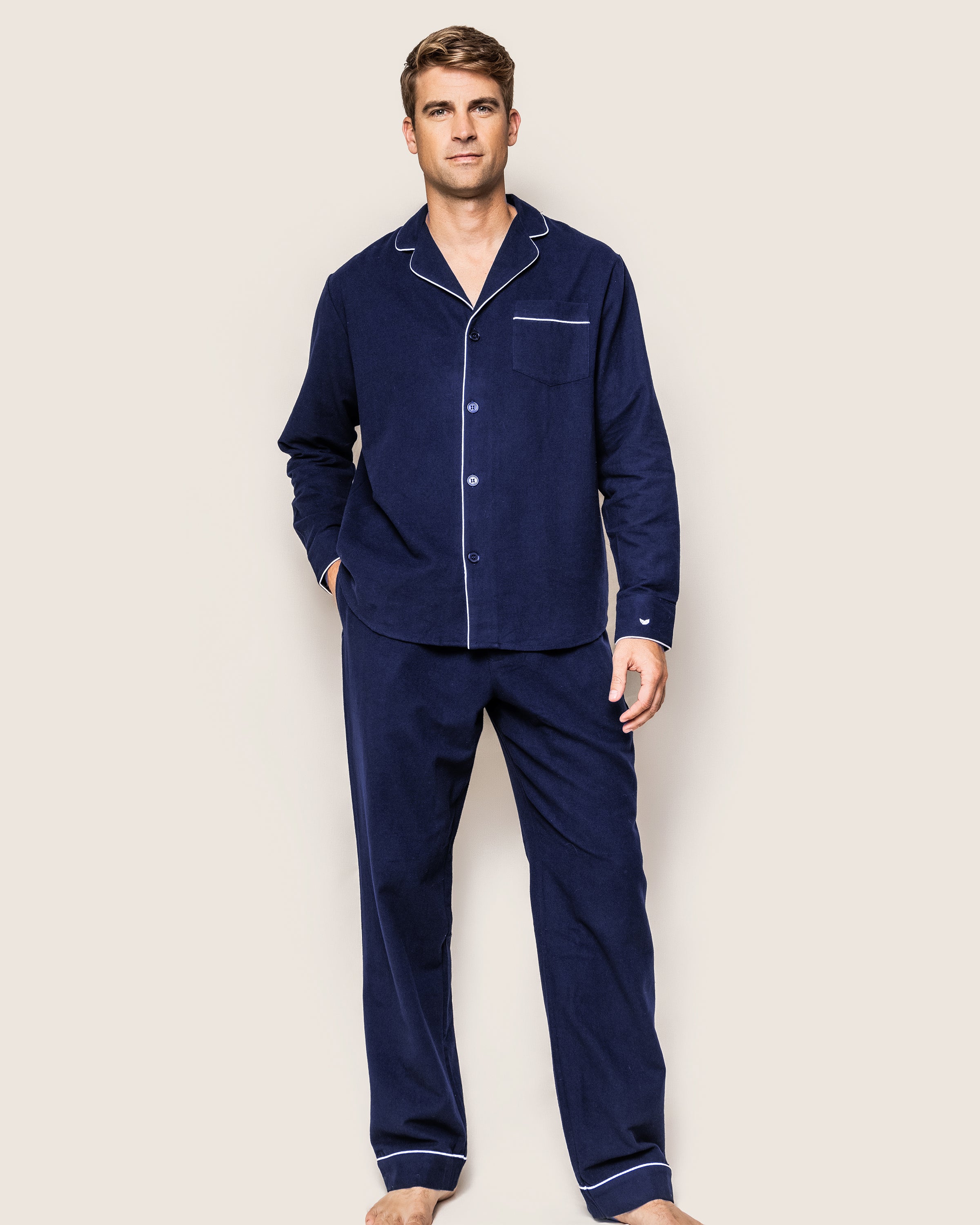 Against a beige backdrop, a man wears the Mens Flannel Pajama Set in Navy by Petite Plume, featuring a collared, button-up top with white piping and matching pants, blending style and comfort.