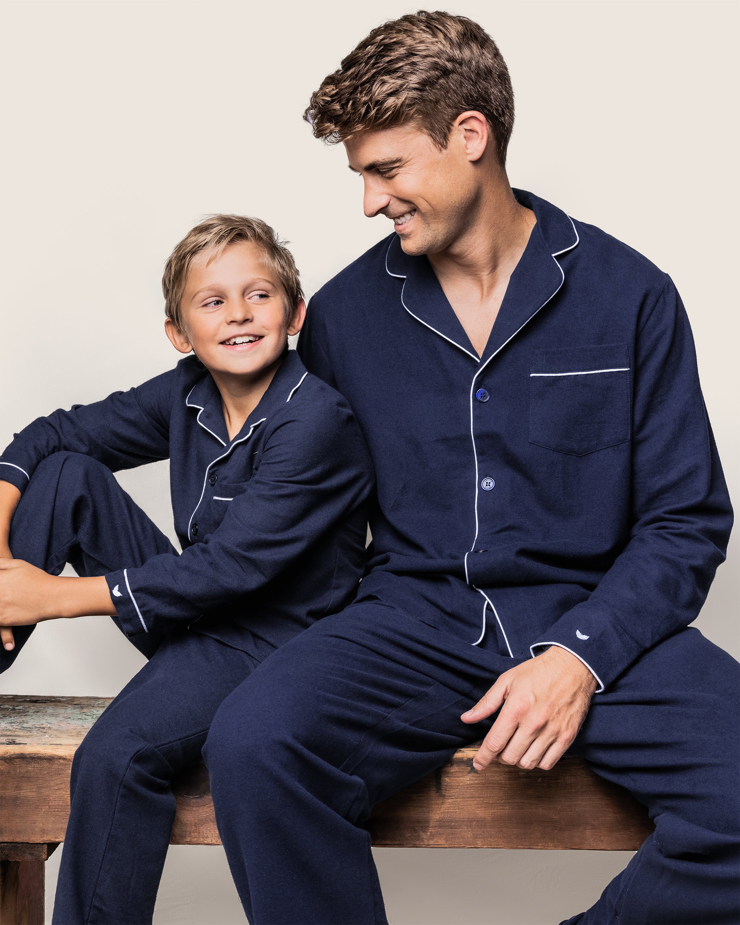 A man and a boy sit on a wooden bench, both in matching Petite Plume Mens Flannel Pajama Sets in navy with white piping. The boy smiles up at the man, who smiles back, against a plain light gray background.