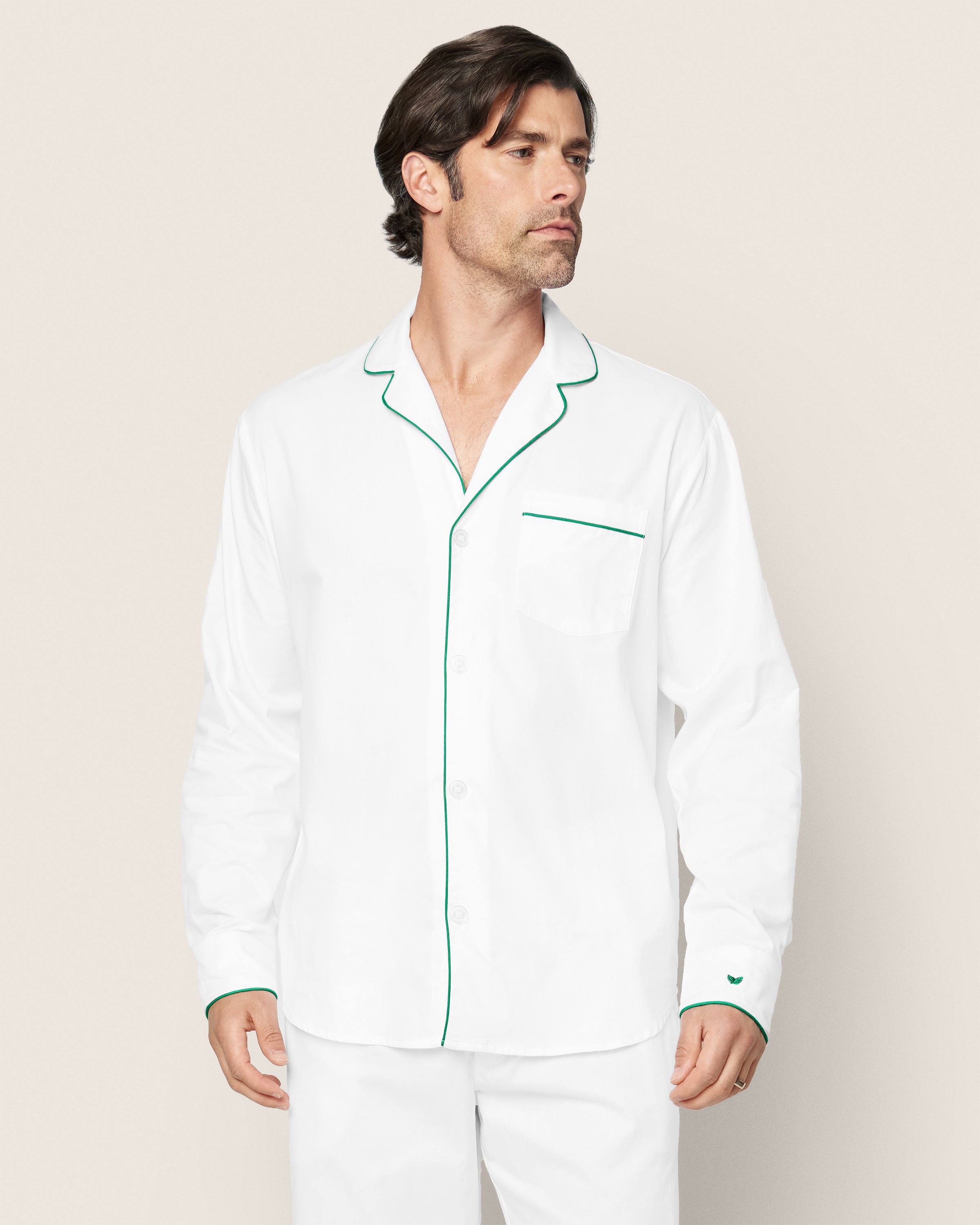 A man models the Petite Plume Mens Twill Pajama Set, featuring a white top with green piping on the collar, cuffs, and pocket. Crafted from premium cotton, his brown hair stands out against a plain, light backdrop.