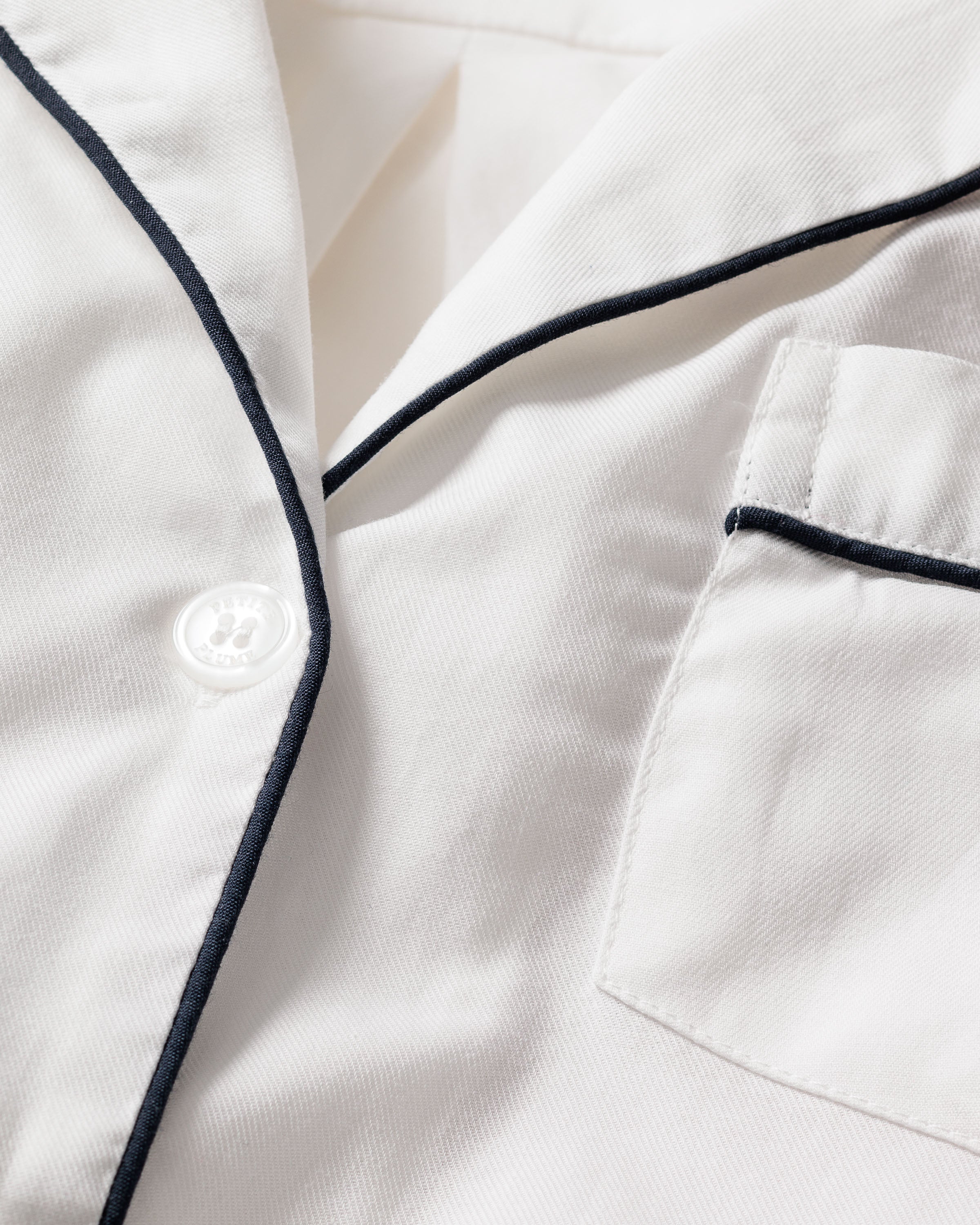 Close-up of a Mens Twill Pajama Set by Petite Plume in white with navy piping, featuring a soft cotton fabric, white buttons, and a neatly tailored chest pocket.