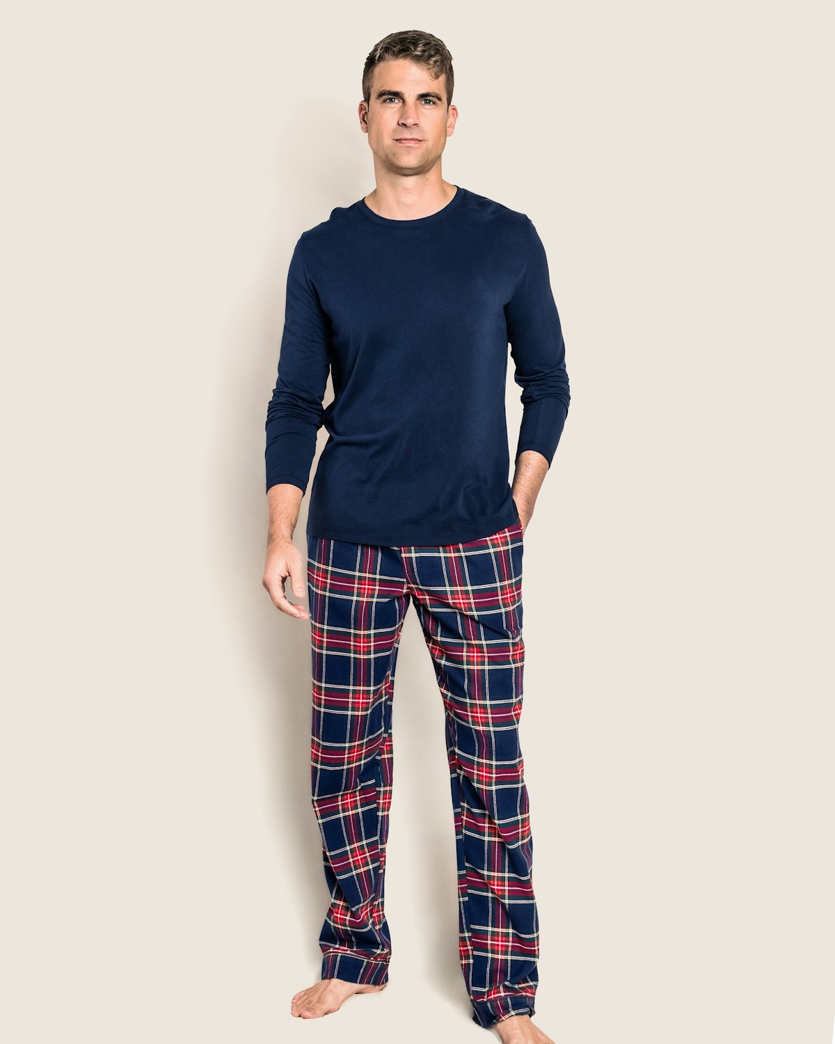 Against a plain background, a person casually stands barefoot with one hand in their pocket, wearing navy long-sleeve shirt and Petite Plumes Men’s Brushed Cotton Tartan Pajama Pant Duo in soft red, blue, and white print.